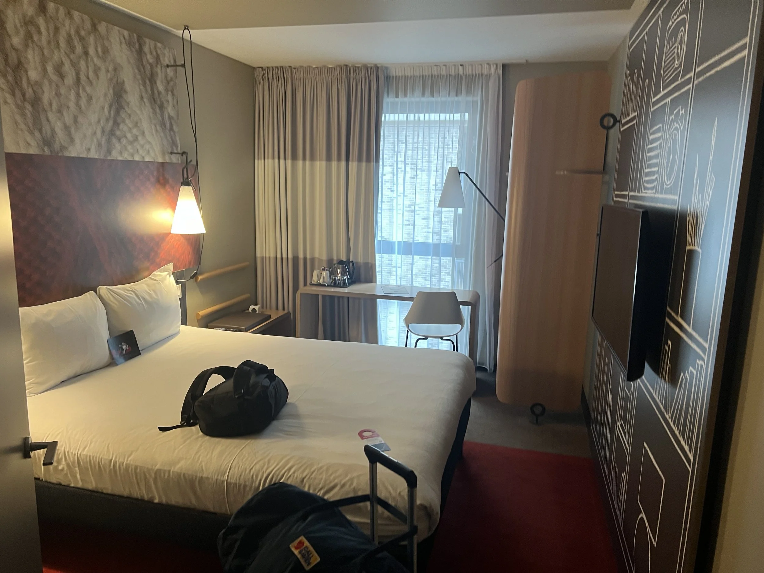 Interior of a hotel room with a bed, nightstand, lamp, window with beige curtains, and a workspace with a chair and table. There is a black backpack on the bed.