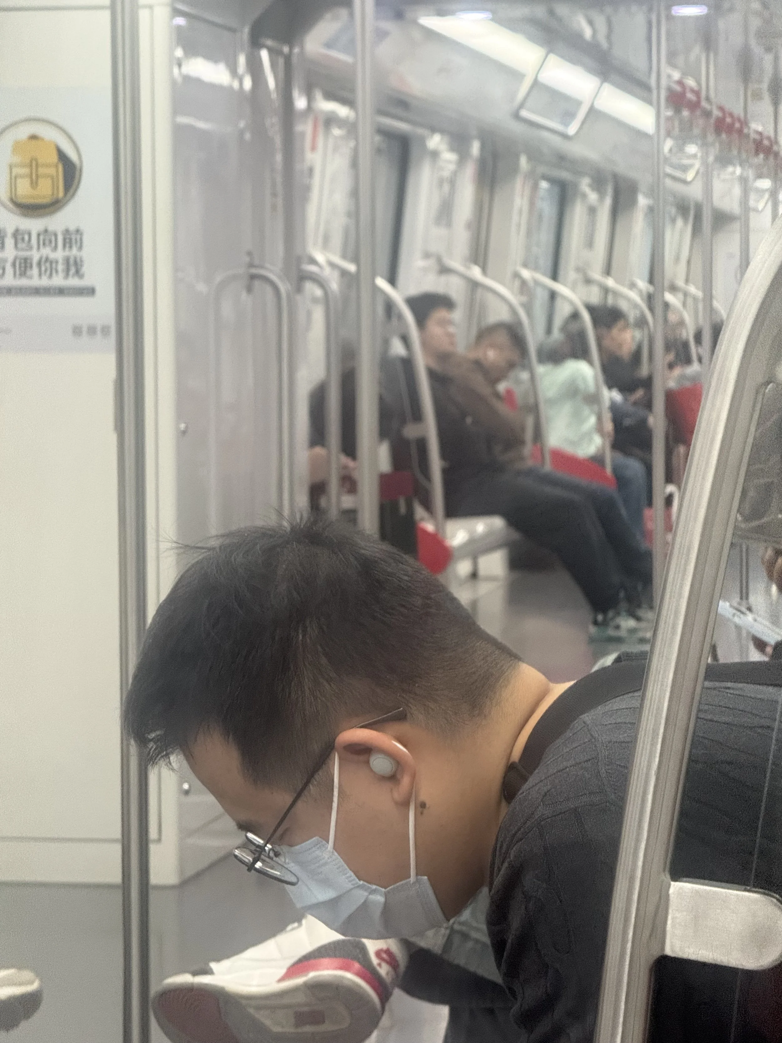 A young man wearing glasses, a face mask, and a black shirt is sleeping on a train seat with his head tilted down and eyes closed. He is wearing white earphones and has a young girl resting her head on his shoulder. The background shows other passeng