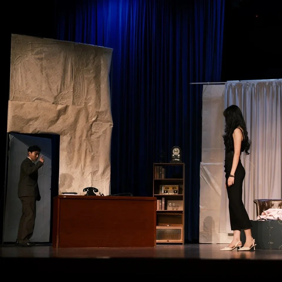 A theater stage scene with a woman in a black dress and heels standing on the right and a man in a suit on the left near a doorway, with a desk, bookshelf, and curtains in the background.