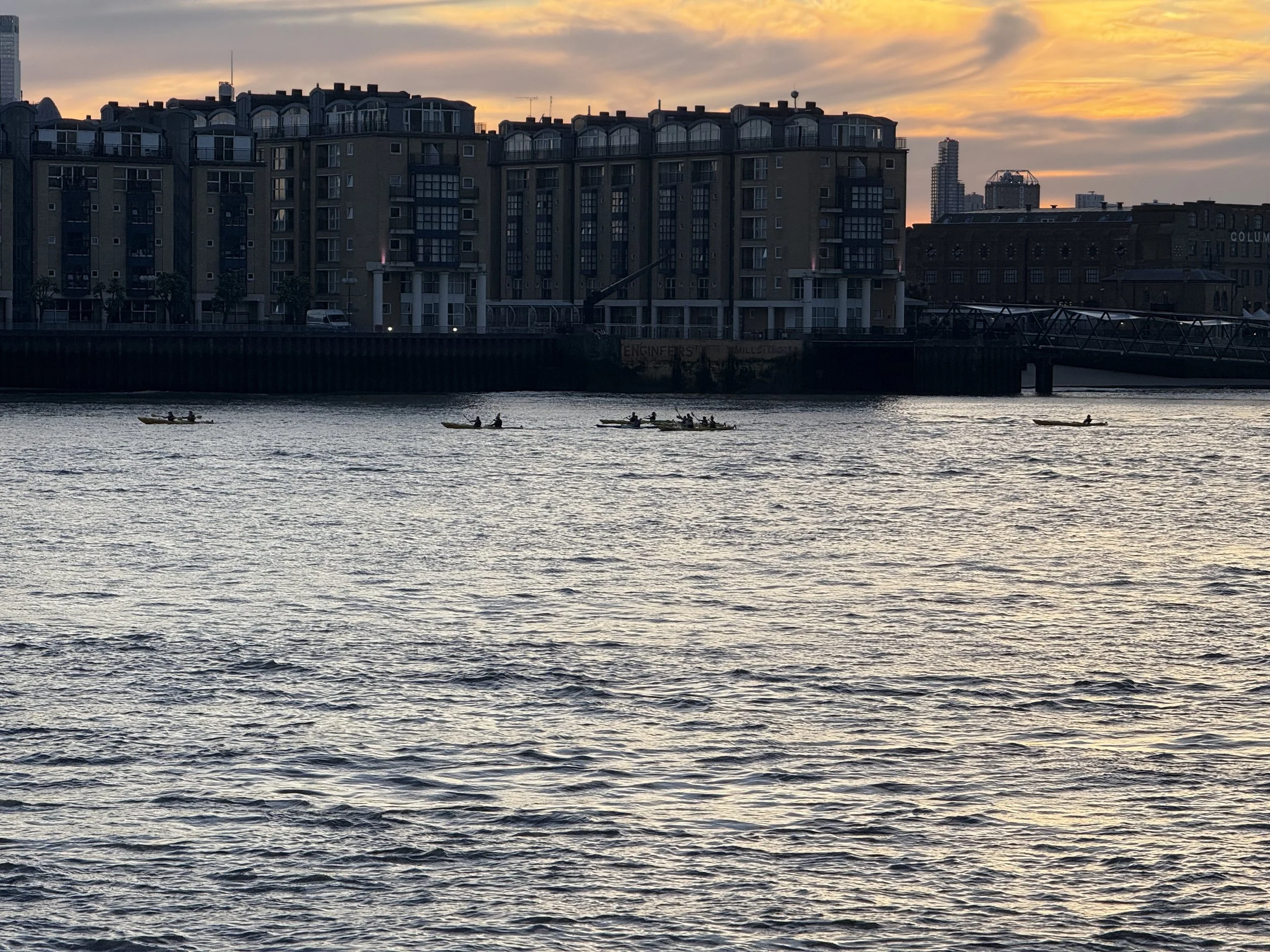 A city skyline at sunset with several kayakers paddling on a river in the foreground.