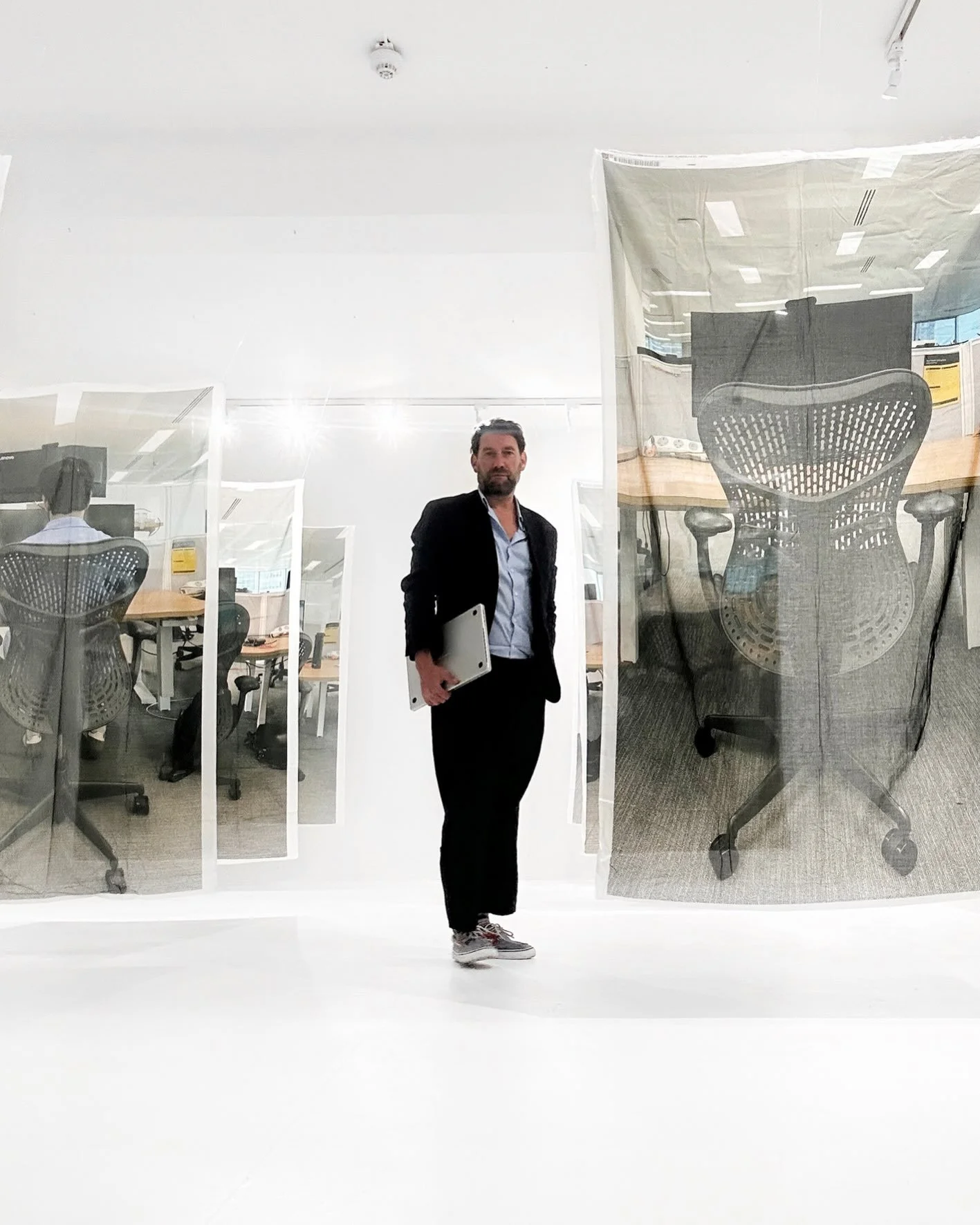 A man wearing a black blazer, blue shirt, and sneakers holding a silver laptop, standing in a modern office with transparent glass walls and office chairs visible behind him.