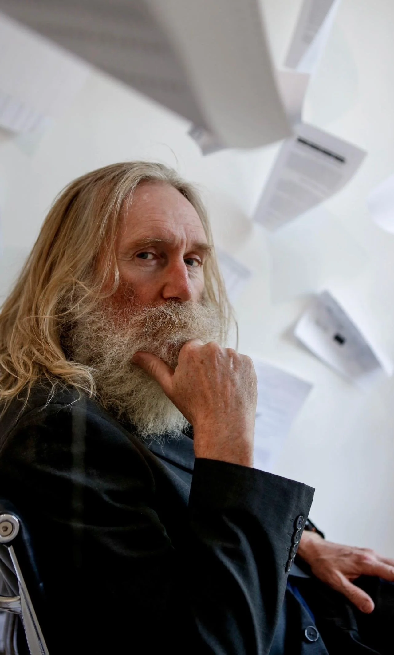 A man with long blonde hair and a beard, sitting indoors. He appears to be deep in thought, with his hand resting on his chin.