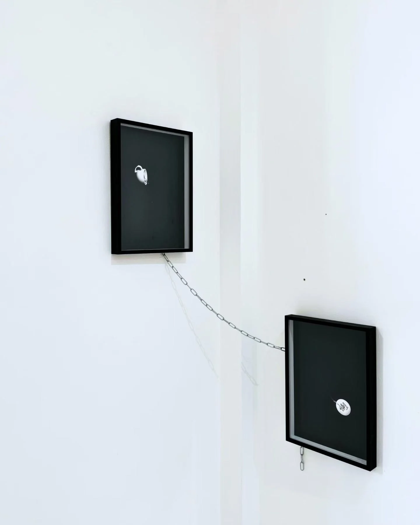 Minimalist art installation with two black framed screens on white walls, connected by a silver chain, each with a modest graphic in the center.