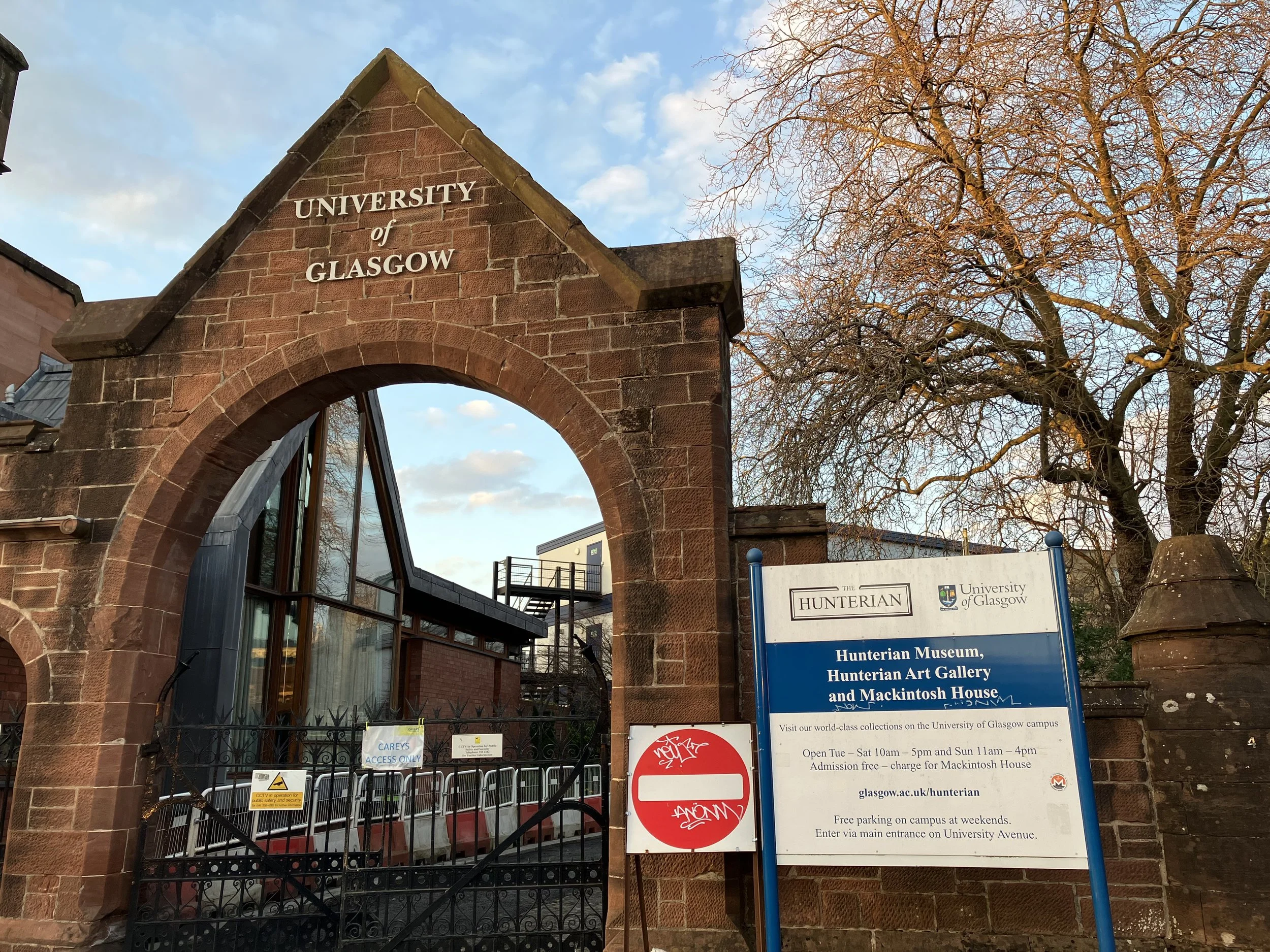 The entrance gate of the University of Glasgow made of red brick with a sign that reads 'University of Glasgow.' There is a large tree without leaves and a blue sky with scattered clouds in the background. A prominent blue informational sign provides
