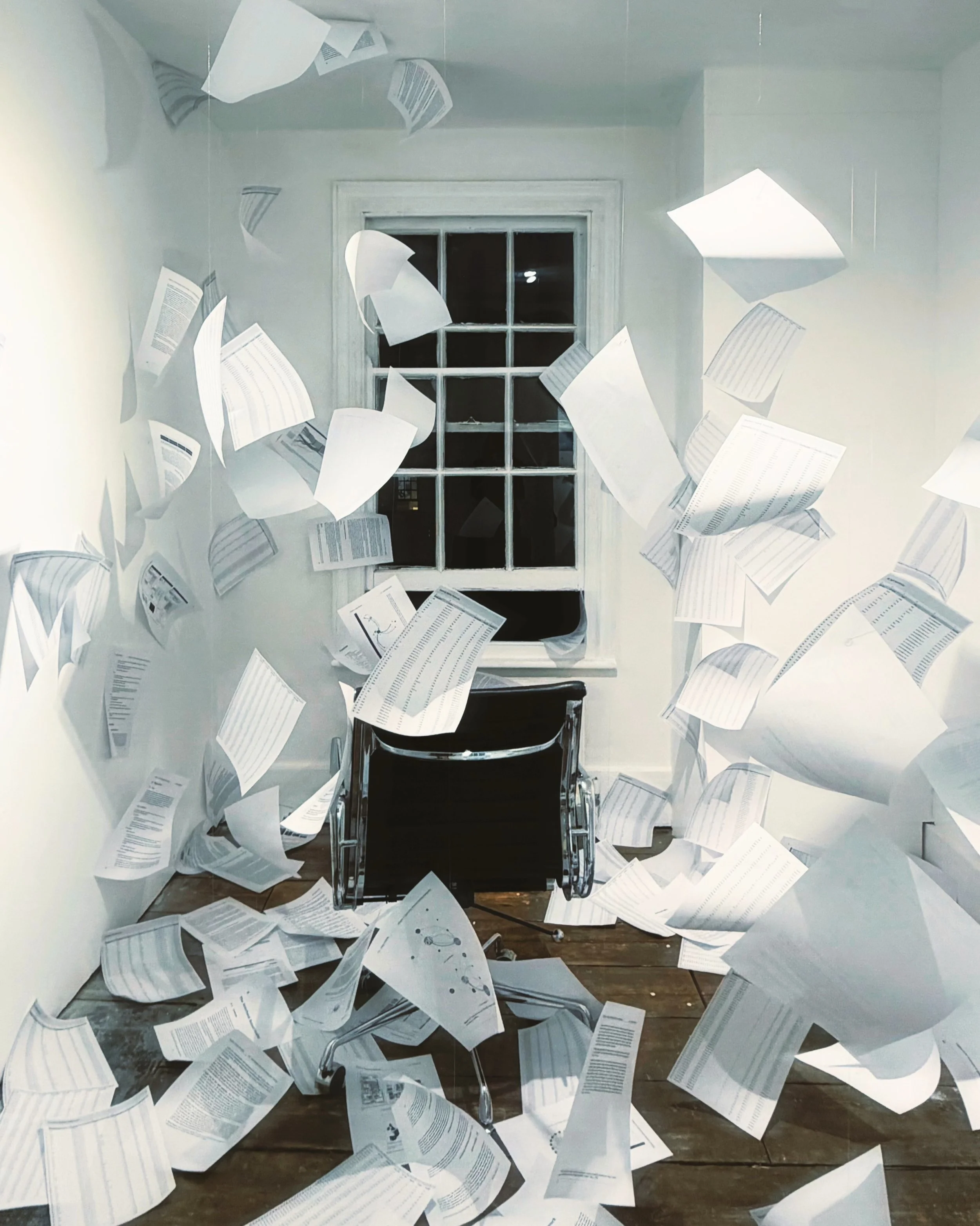 A room with a large window at the back, papers flying around and scattered on the floor, four-wheel chair in the middle, and white walls.