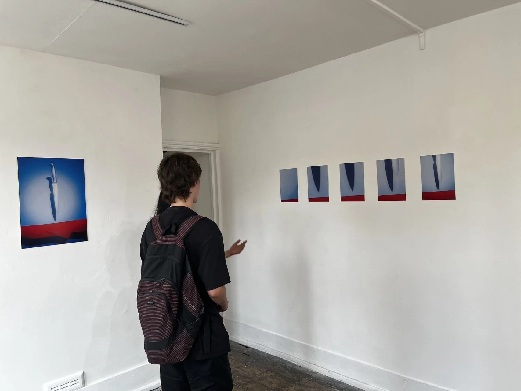 An art gallery with five paintings on the wall depicting a blue and red abstract figure, and a person with brown hair, wearing a black shirt and a black and red backpack, observing the artwork.