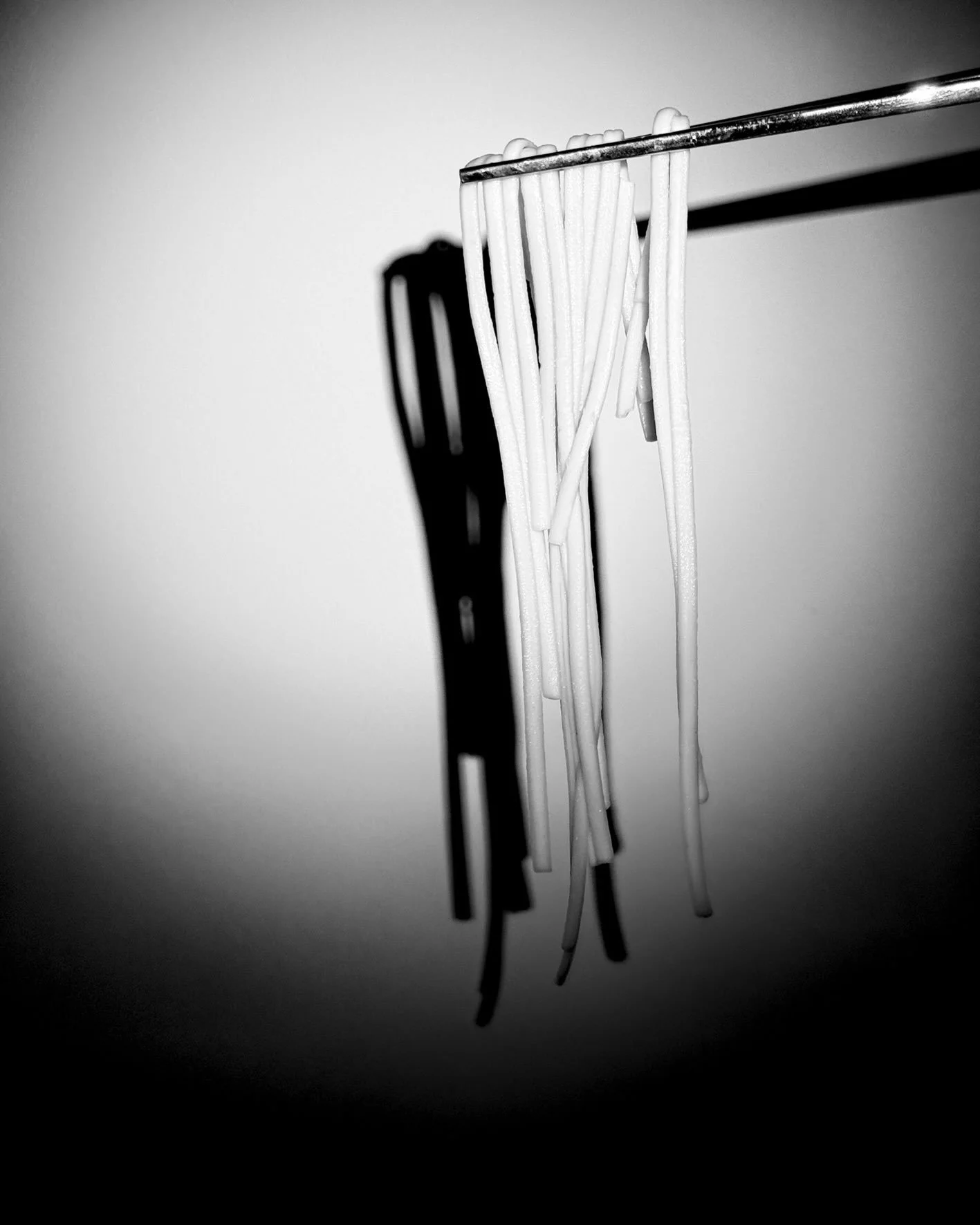 Black and white photograph of a bundle of uncooked spaghetti hanging from a metal rod, casting a shadow on the wall behind.