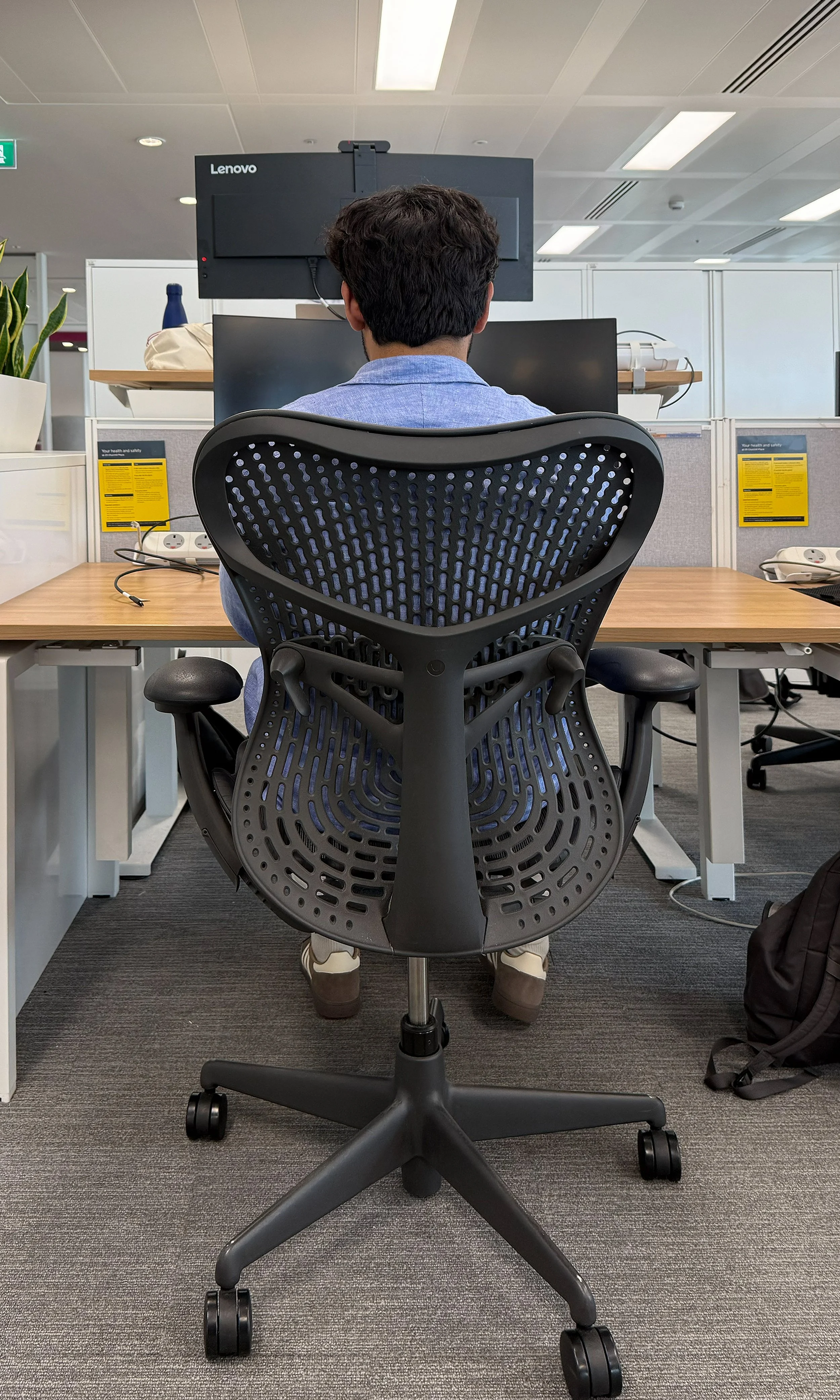 Person sitting at a desk in an office workspace, viewed from behind, using a computer with a large monitor, surrounded by office equipment and supplies.
