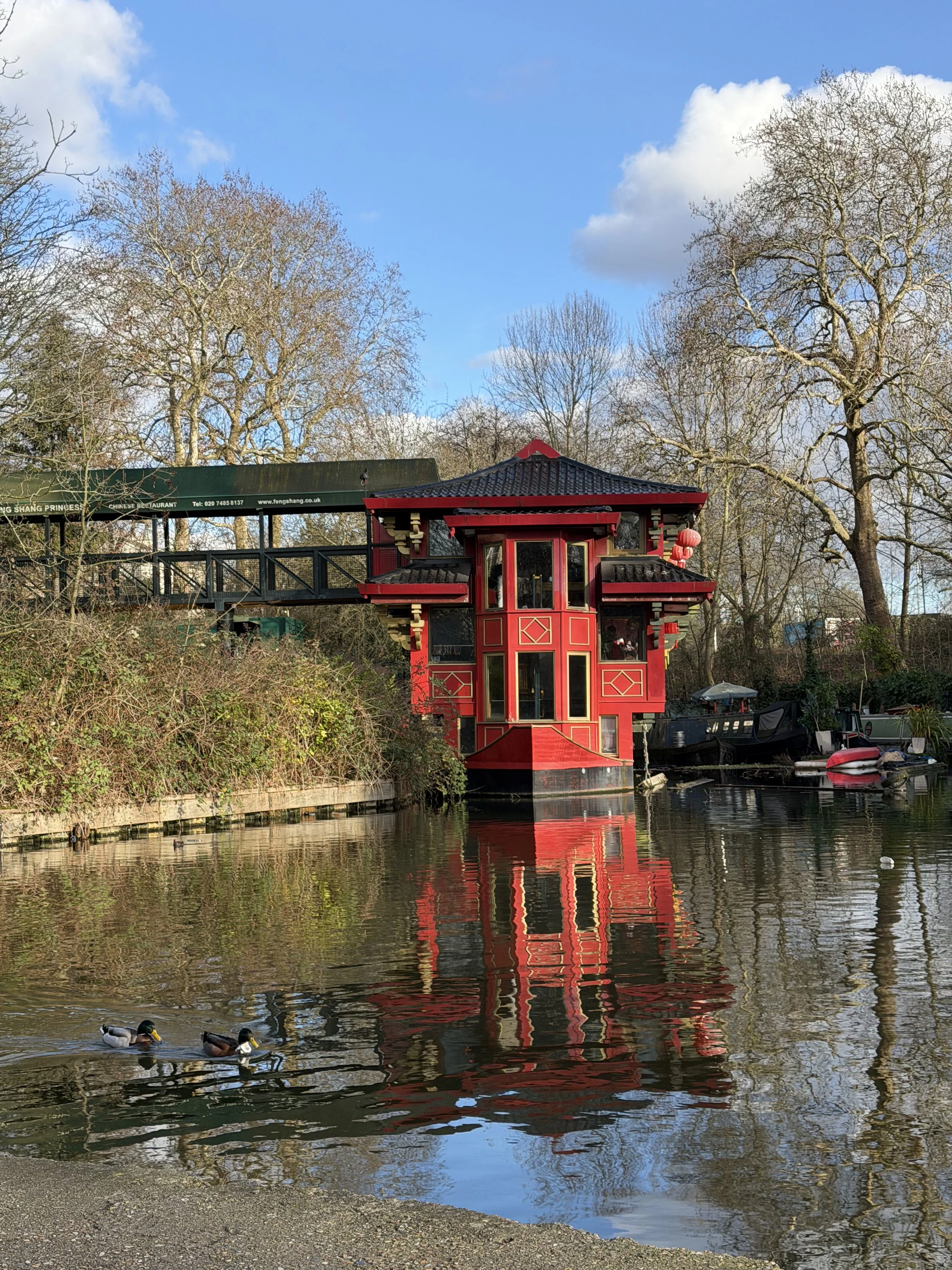 A small red and black boat house on the water with ducks swimming nearby. Behind it, there are leafless trees and a building with a green sign that has Chinese characters and a website address. The sky is partly cloudy with blue patches visible.