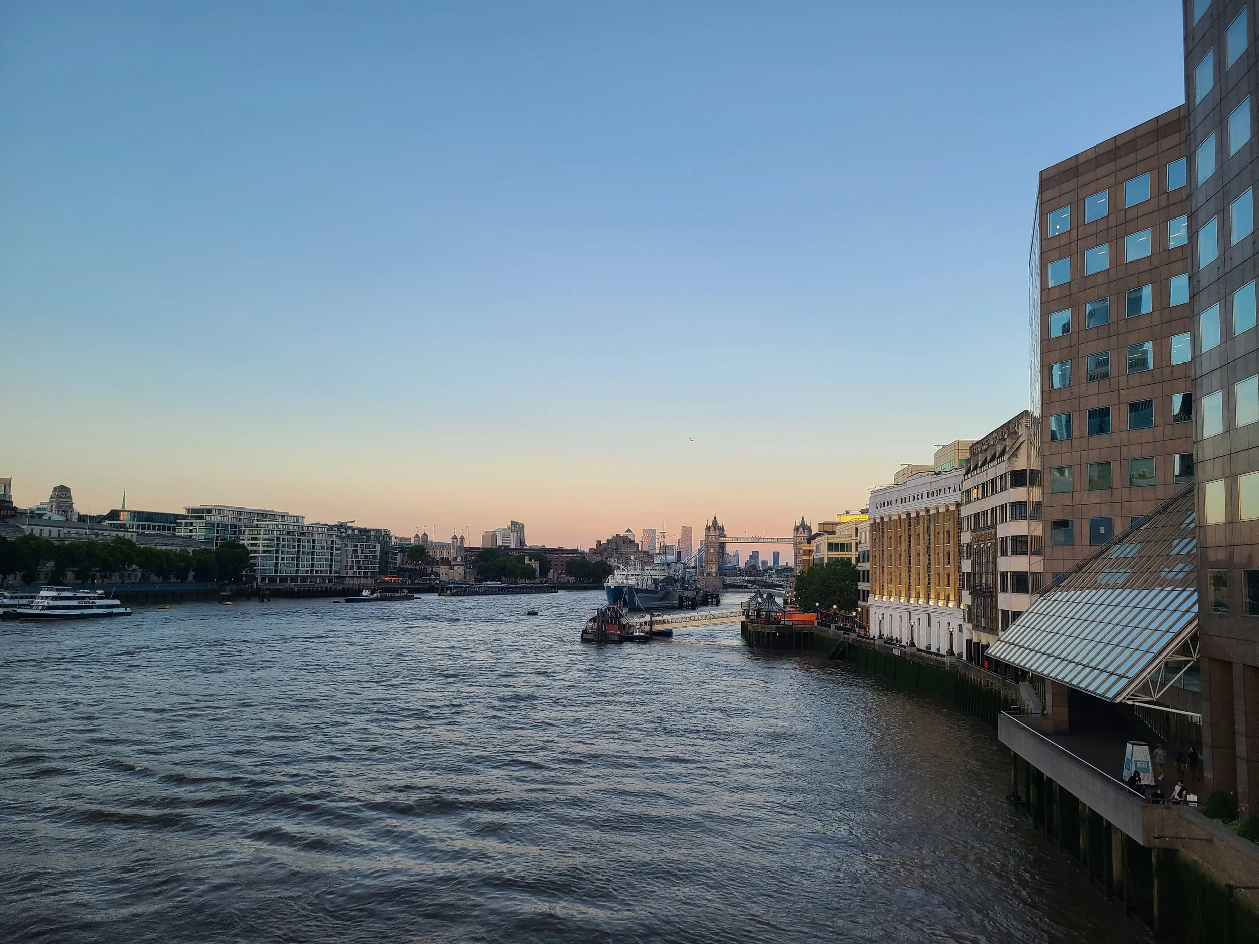 A cityscape view over a river with modern buildings on the right side, including a building with many windows and a stone facade, and historical structures with towers in the background. The sky is clear with a slight gradient of light blue and the r