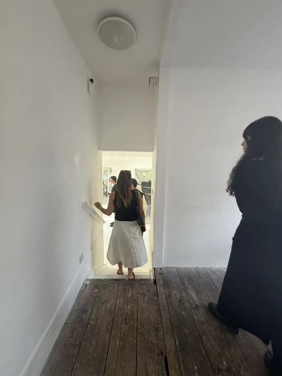 A woman in a black top and white skirt descending a staircase with her back to the camera, while another woman in black attire stands on the wooden floor to the right. The staircase is lit by natural light from the door at the bottom, with white wall
