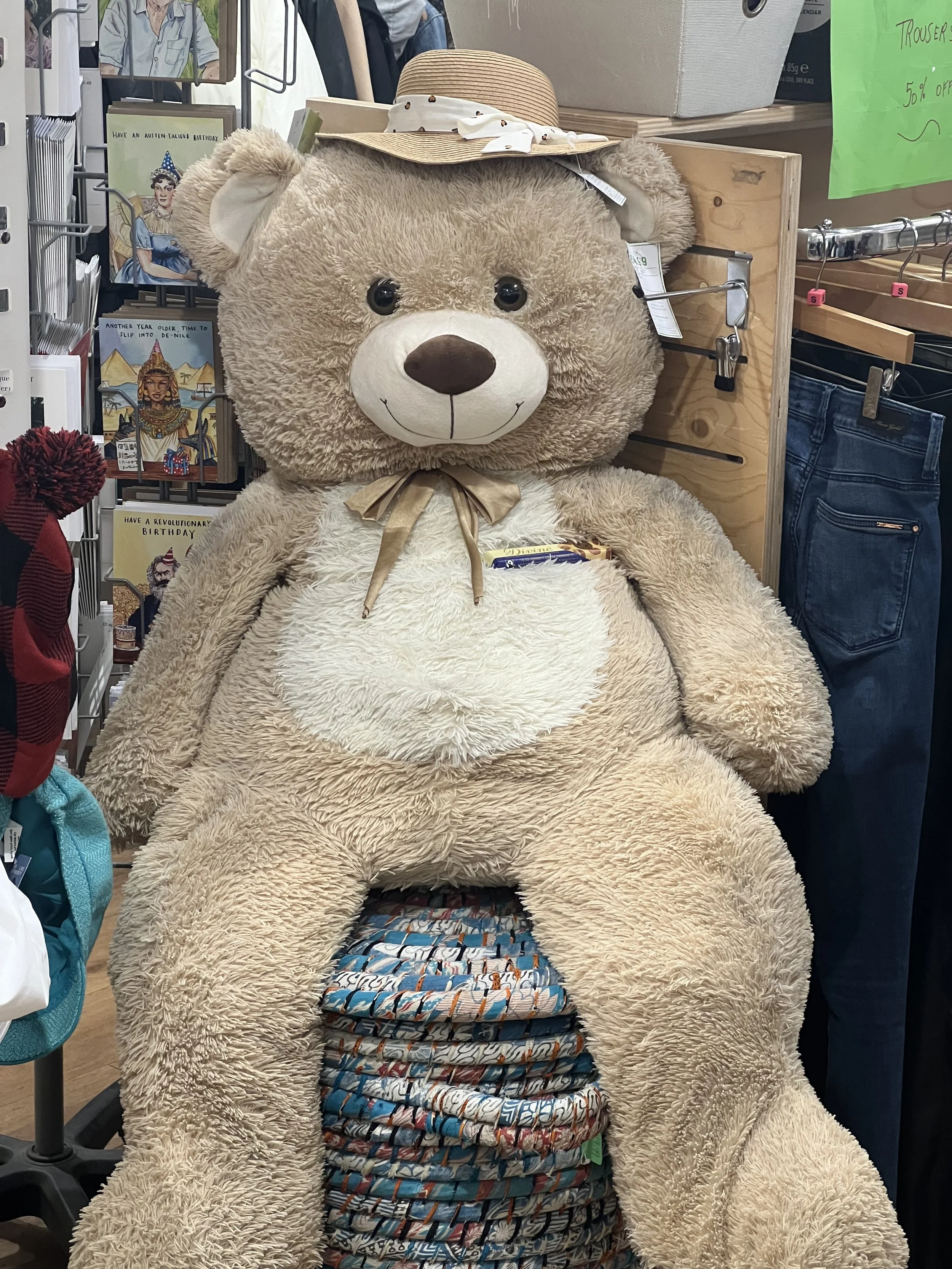 A large teddy bear with beige fur and a white snout, wearing a straw hat with a white bow, sitting among folded cloth items or blankets with various patterns in a store. Behind it are shelves with greeting cards, books, and clothing.