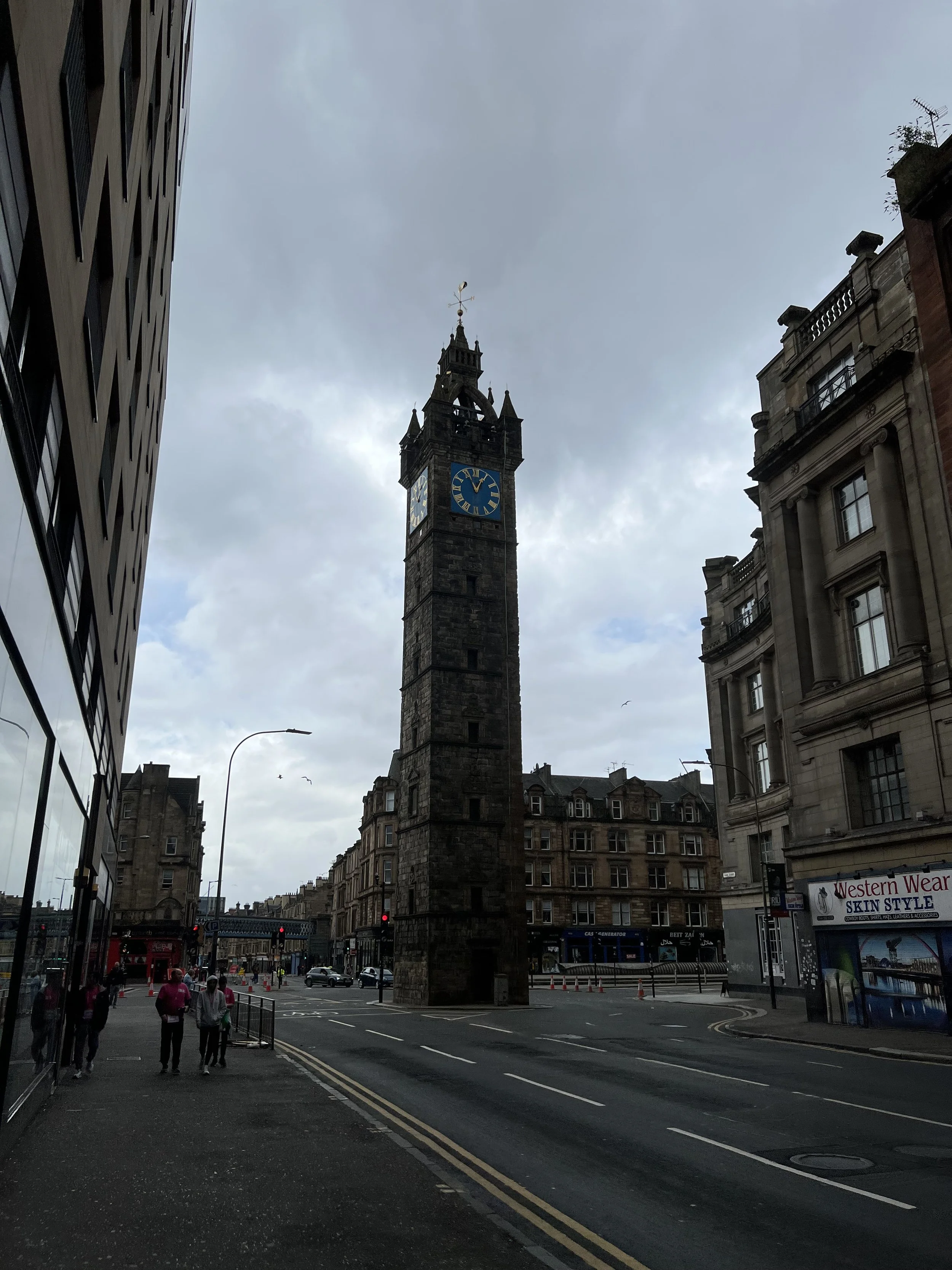 Street view with a historic clock tower in the center, surrounded by modern and historic buildings, cloudy sky overhead, people walking on the sidewalk, and traffic on the road.