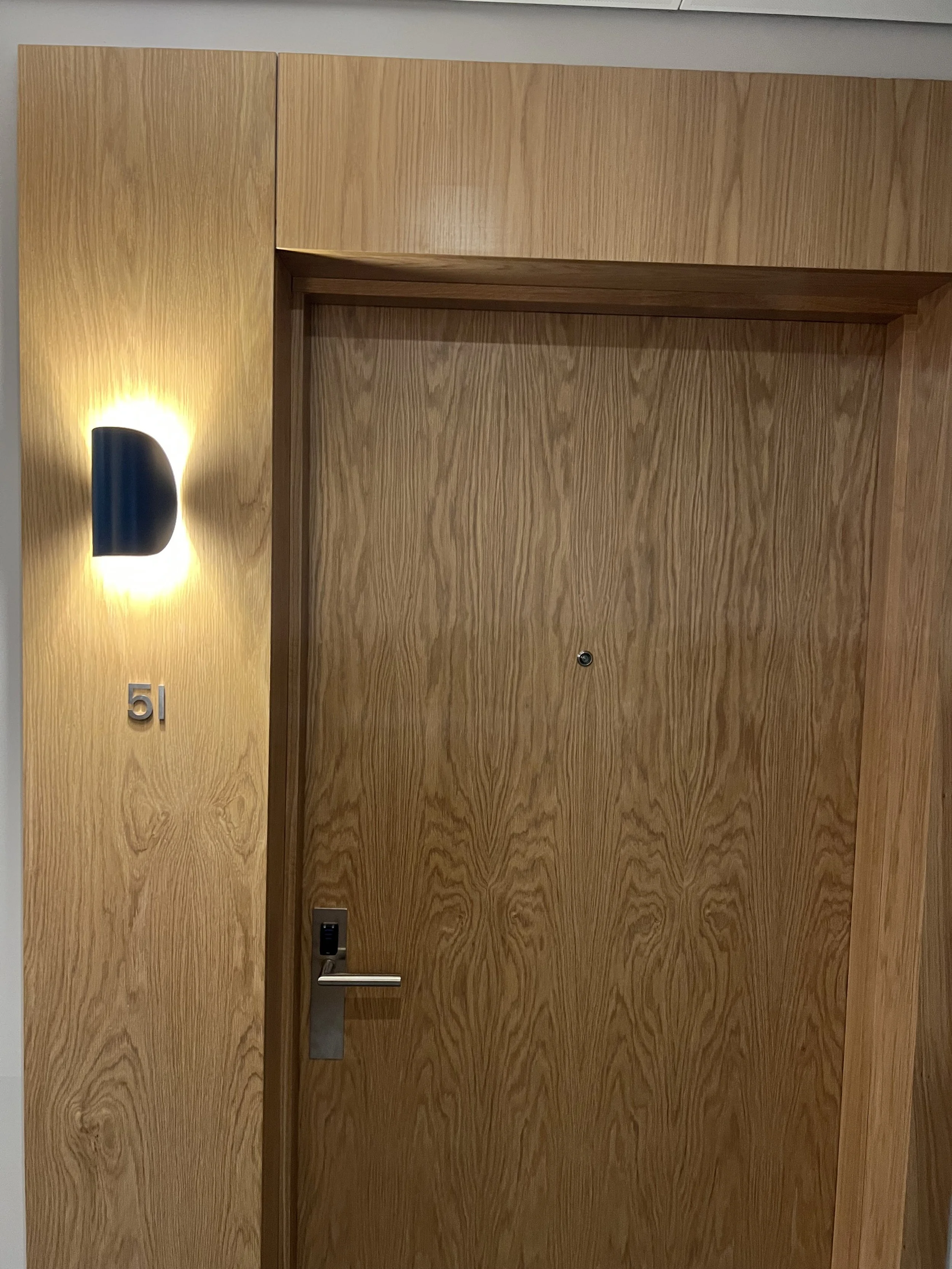A wooden door with a metallic handle, a peephole, a black lock at the bottom, a black doorbell, and a metallic number '51' on the left side. A wall-mounted lamp is on the left side, illuminating the area.