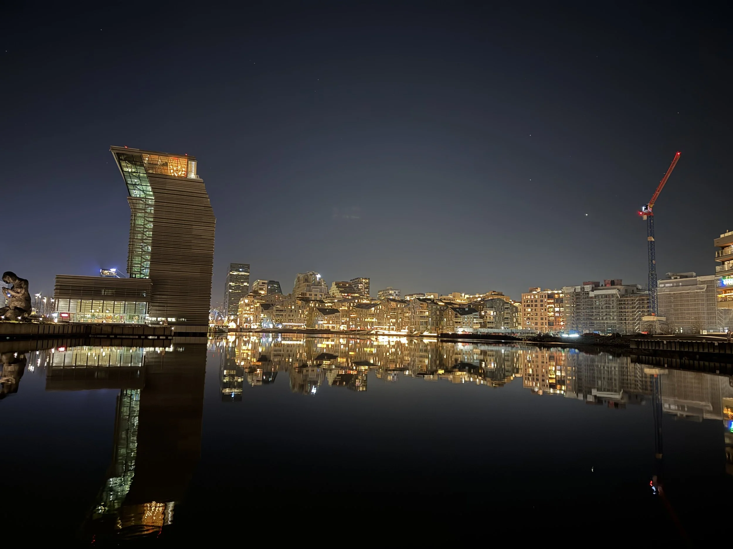 Night cityscape featuring modern buildings, cranes, and reflected city lights on calm water.