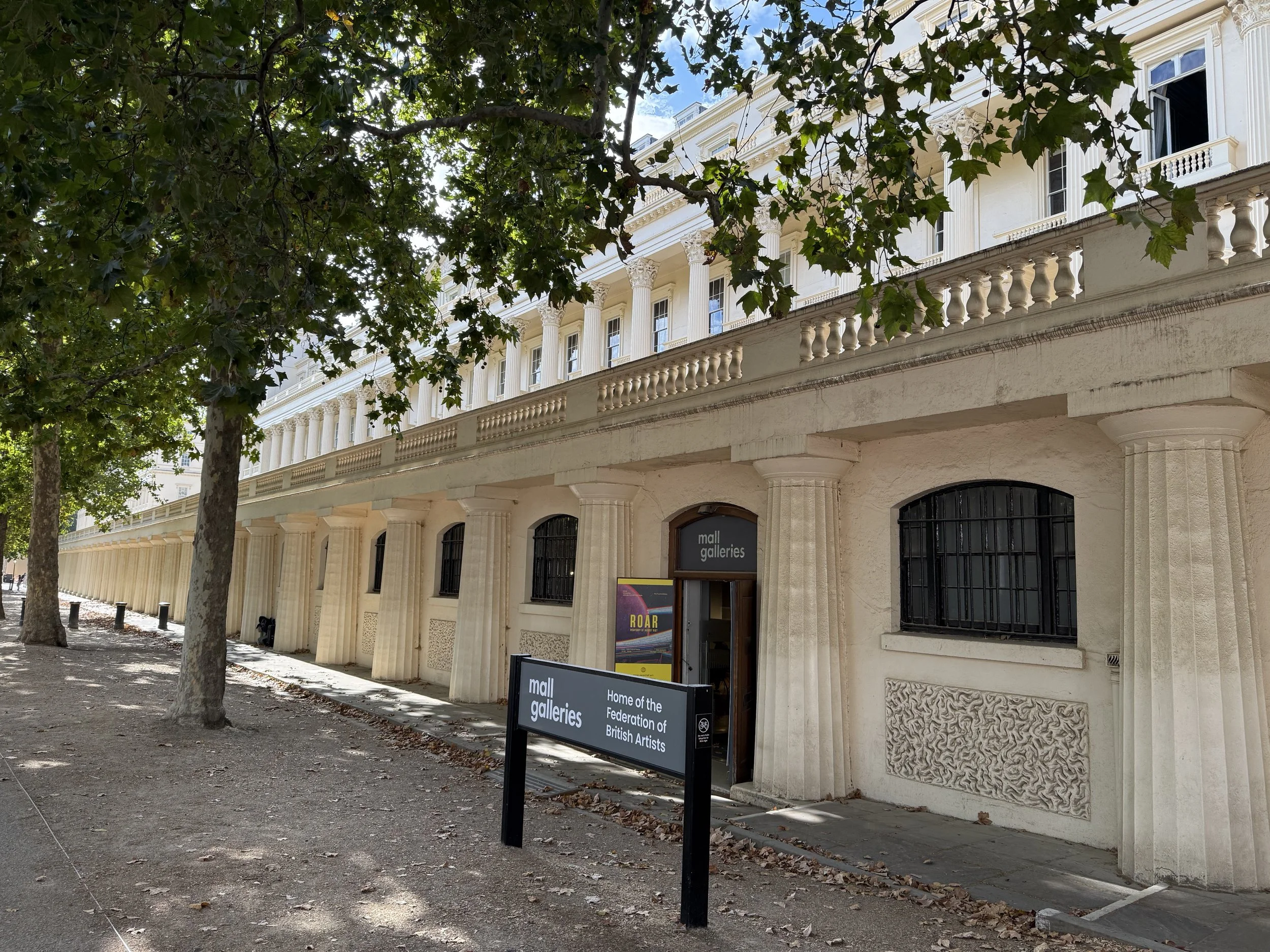 View of a historic building with classical columns and a white facade, surrounded by trees with green leaves, and a sign in front indicating it is the mall galleries, home of the Federation of British Artists.