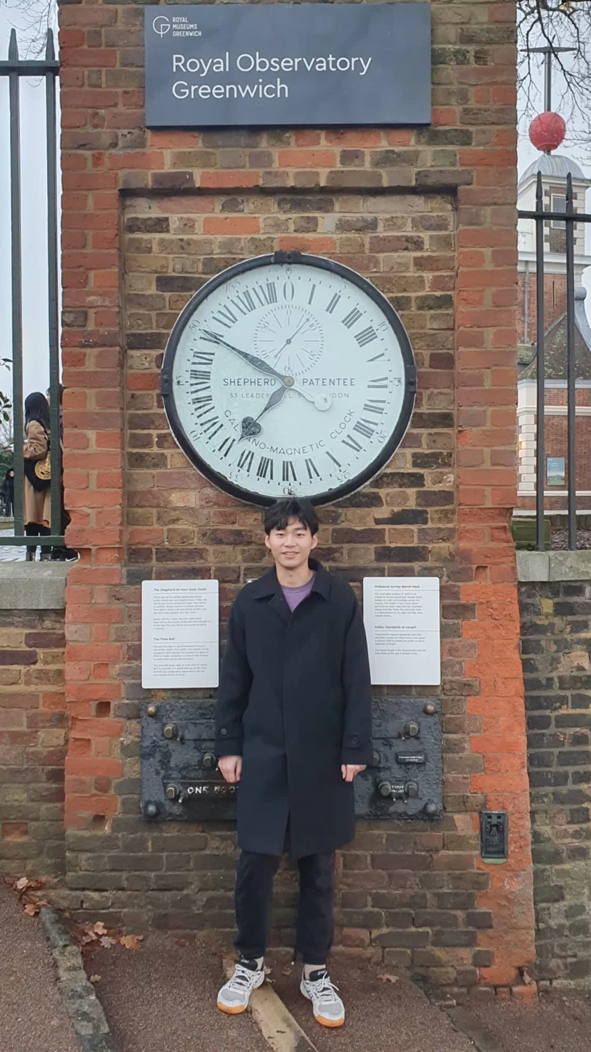 A young man standing in front of a large clock on a brick wall at the Royal Observatory Greenwich. The clock shows the time as approximately 4:50.