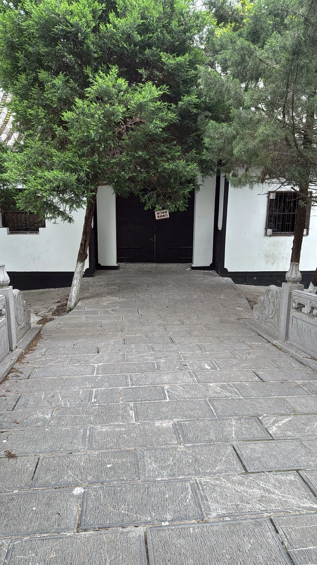 Stone pathway leading to black gate flanked by white walls and green trees.