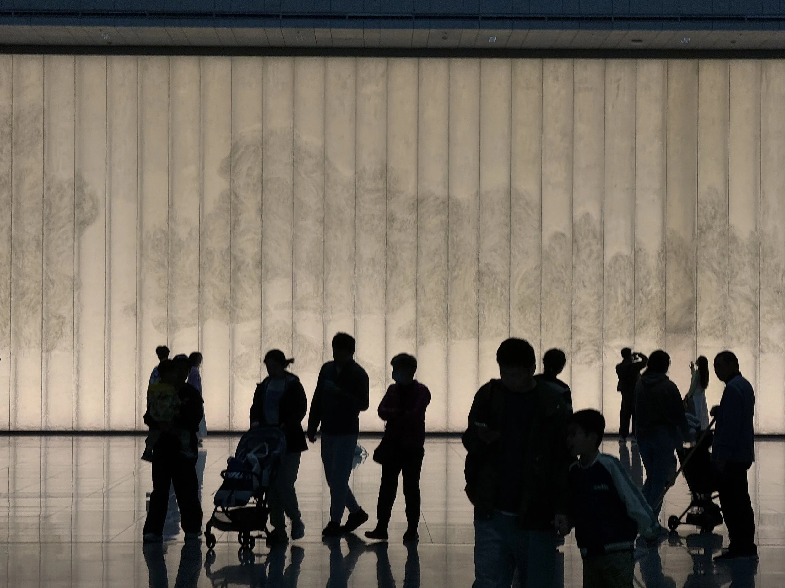 A group of people standing in front of a large illuminated wall with horizontal panels, possibly in an indoor public space.