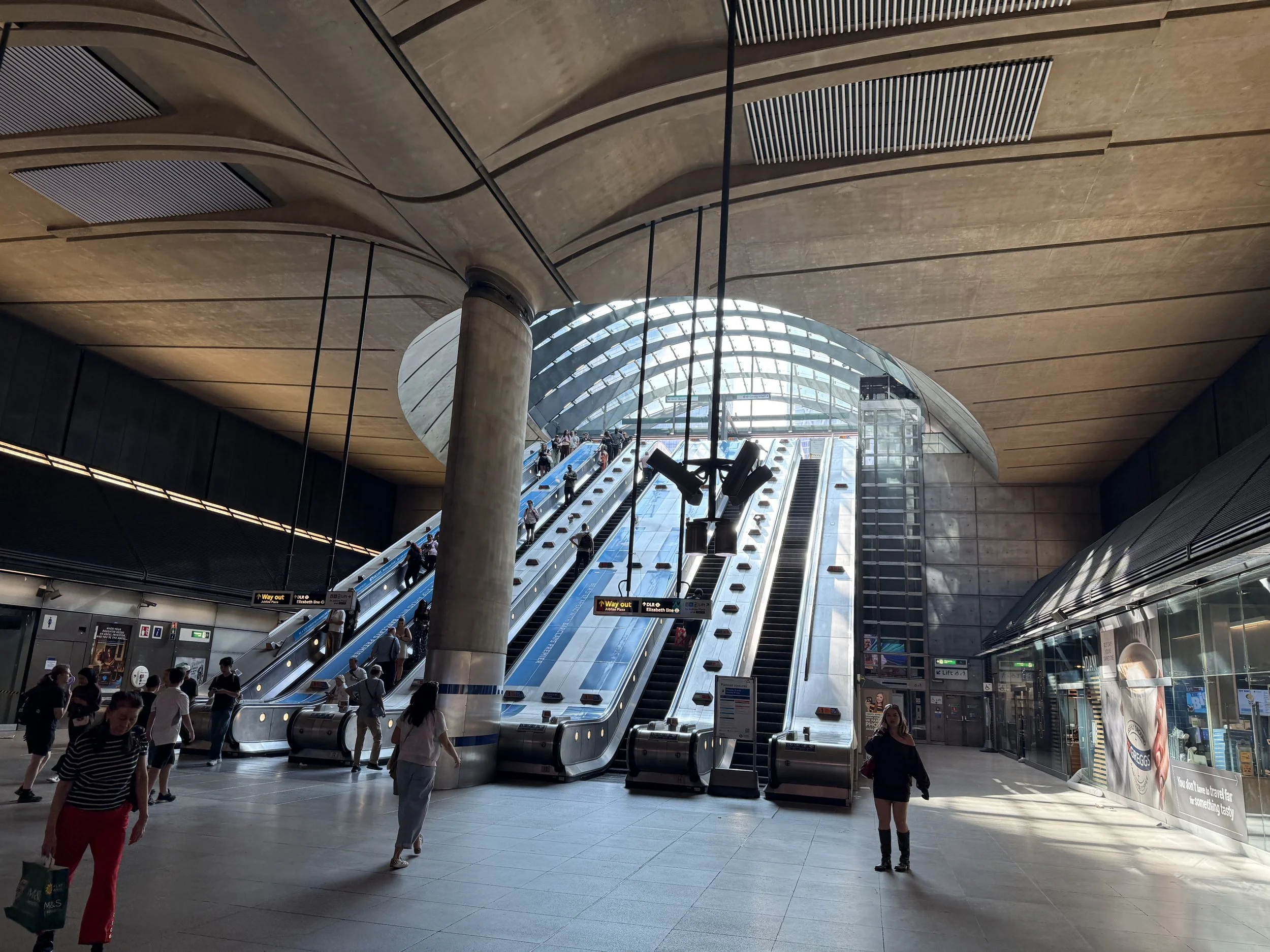 People walking inside a modern train or subway station with escalators going up, a curved glass ceiling, and signage indicating directions.