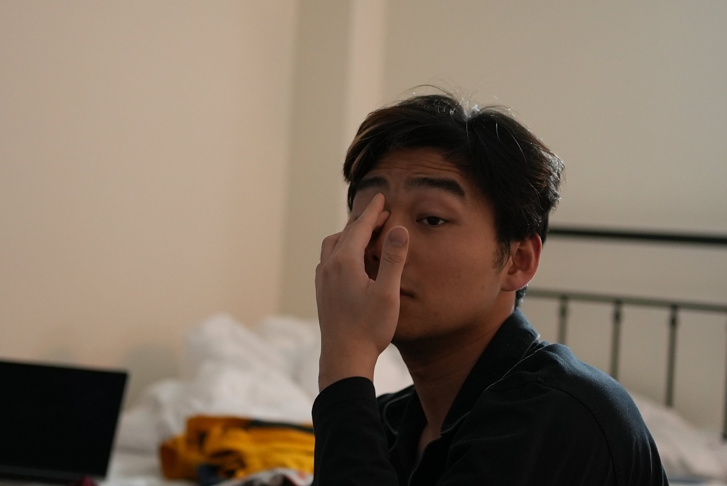 A young man with dark hair and light skin is sitting in a room, touching his forehead with his hand, and looking down. He is wearing a black shirt. The background features a bed and a dresser.