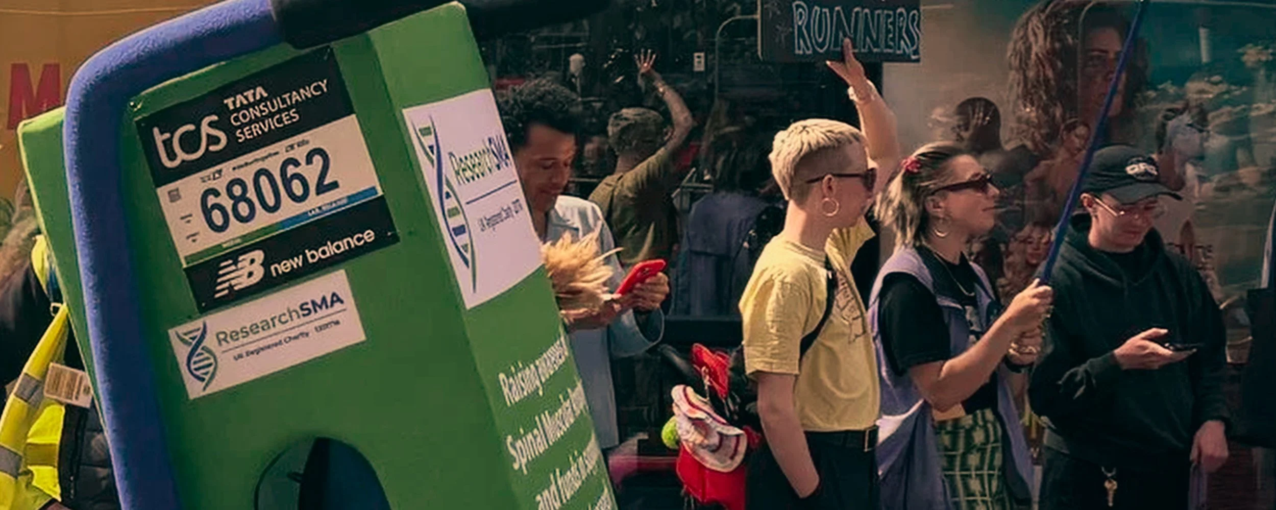A group of people waiting at a bus stop, with some looking at their phones. A large green bus stop sign in the foreground displays logos for TCS, New Balance, ResearchSMA, and other sponsors. Behind the group, a bus with the word 'ROAMERS' on it is v