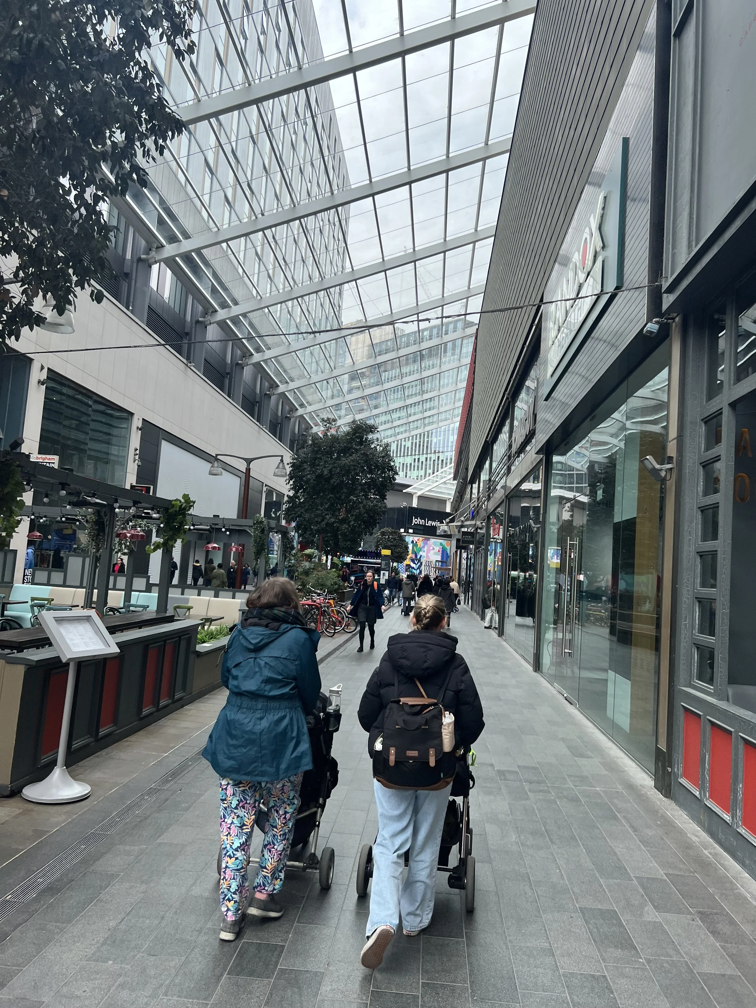 People walking in a shopping mall with a glass ceiling, some pushing strollers and others walking alone, with storefronts on the right and outdoor seating on the left.