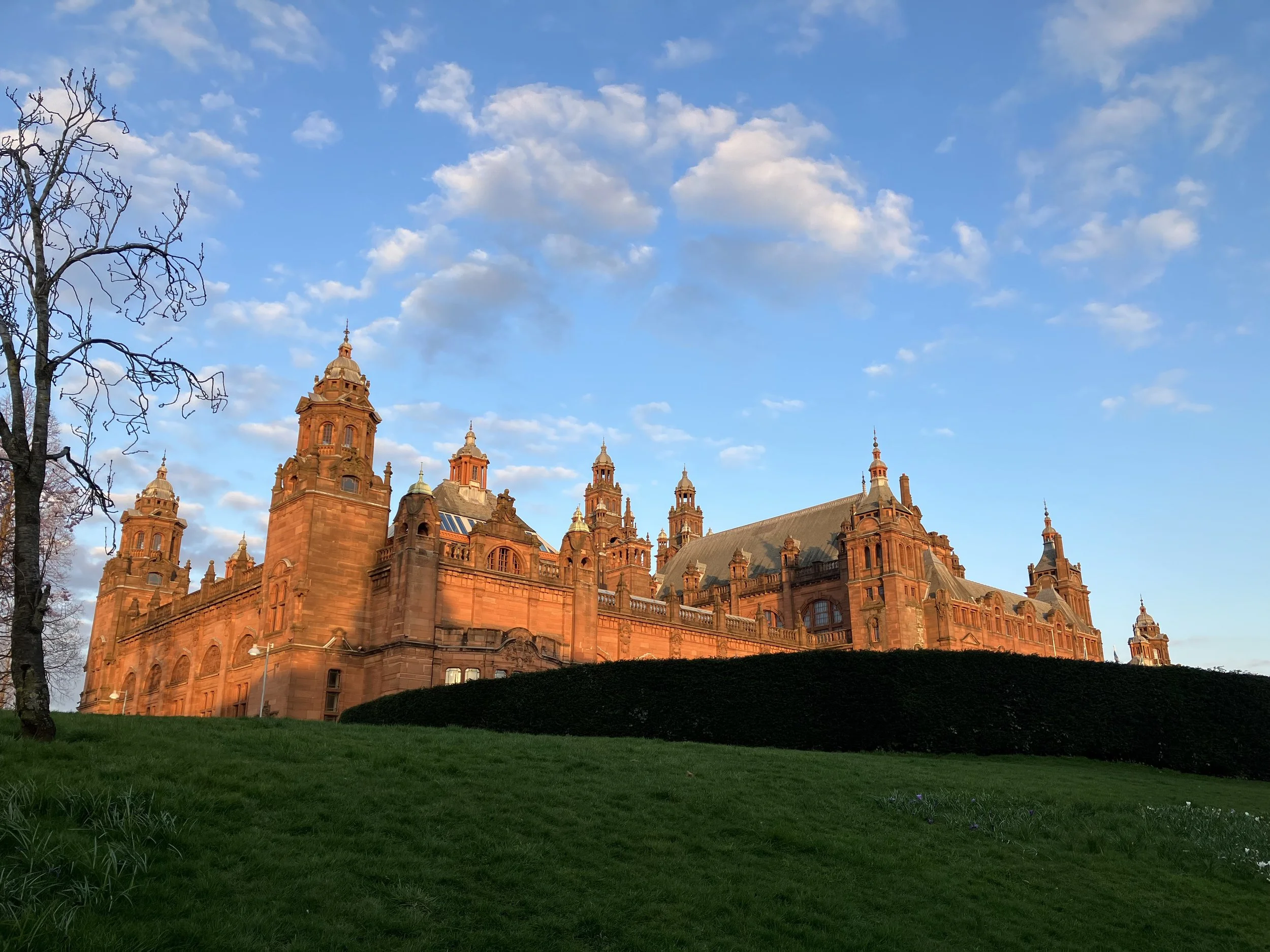 A large historic castle with multiple towers and spires made of red stone, set against a blue sky with scattered clouds. There are leafless trees to the left and a grassy area in the foreground.