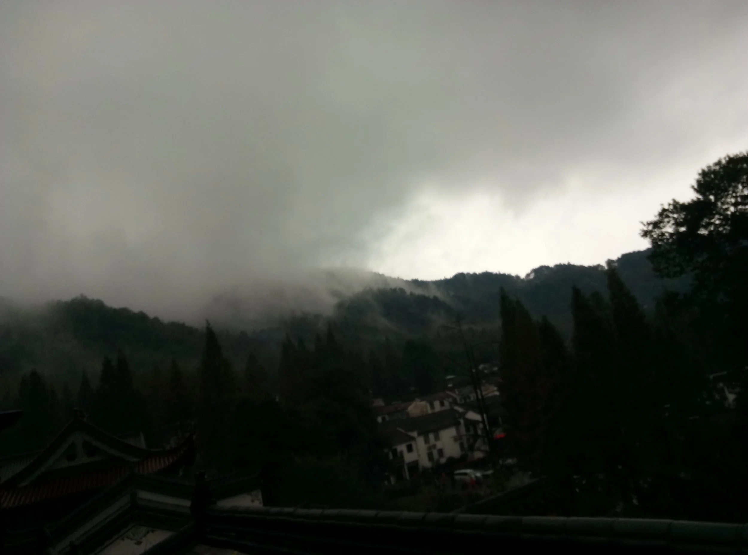 Dark stormy sky with thick gray clouds over a mountainous landscape and a small town or village with rooftops visible at the bottom of the image.