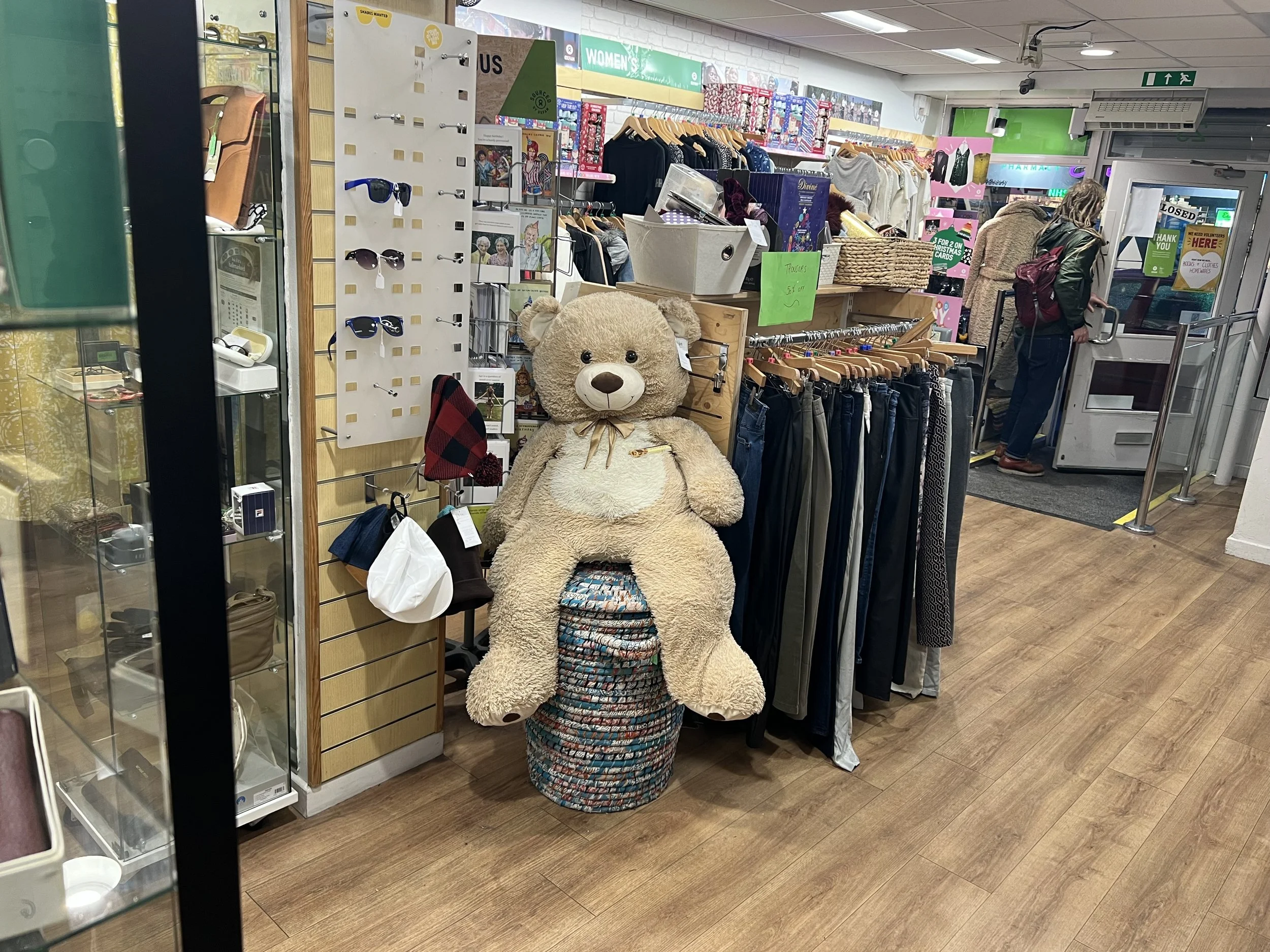 Inside a thrift store, a large teddy bear sitting on a displays stacked with multicolored woven baskets, with clothes racks, sunglasses display, and a customer at the register in the background.