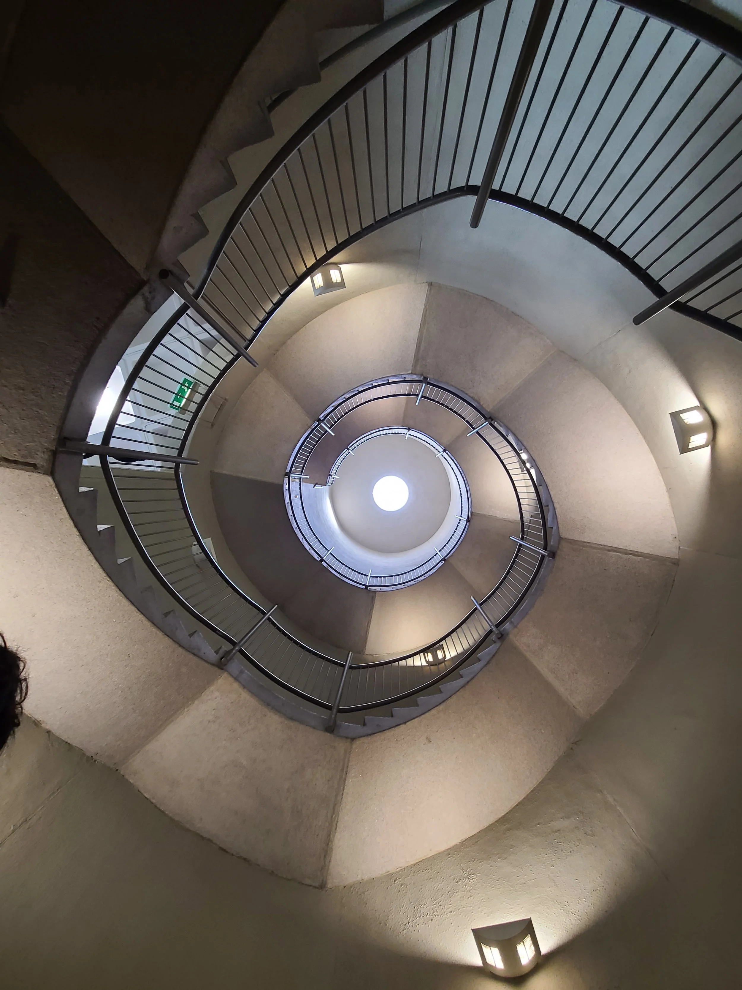 A spiral staircase viewed from below, with metal handrails and light fixtures illuminating the area.