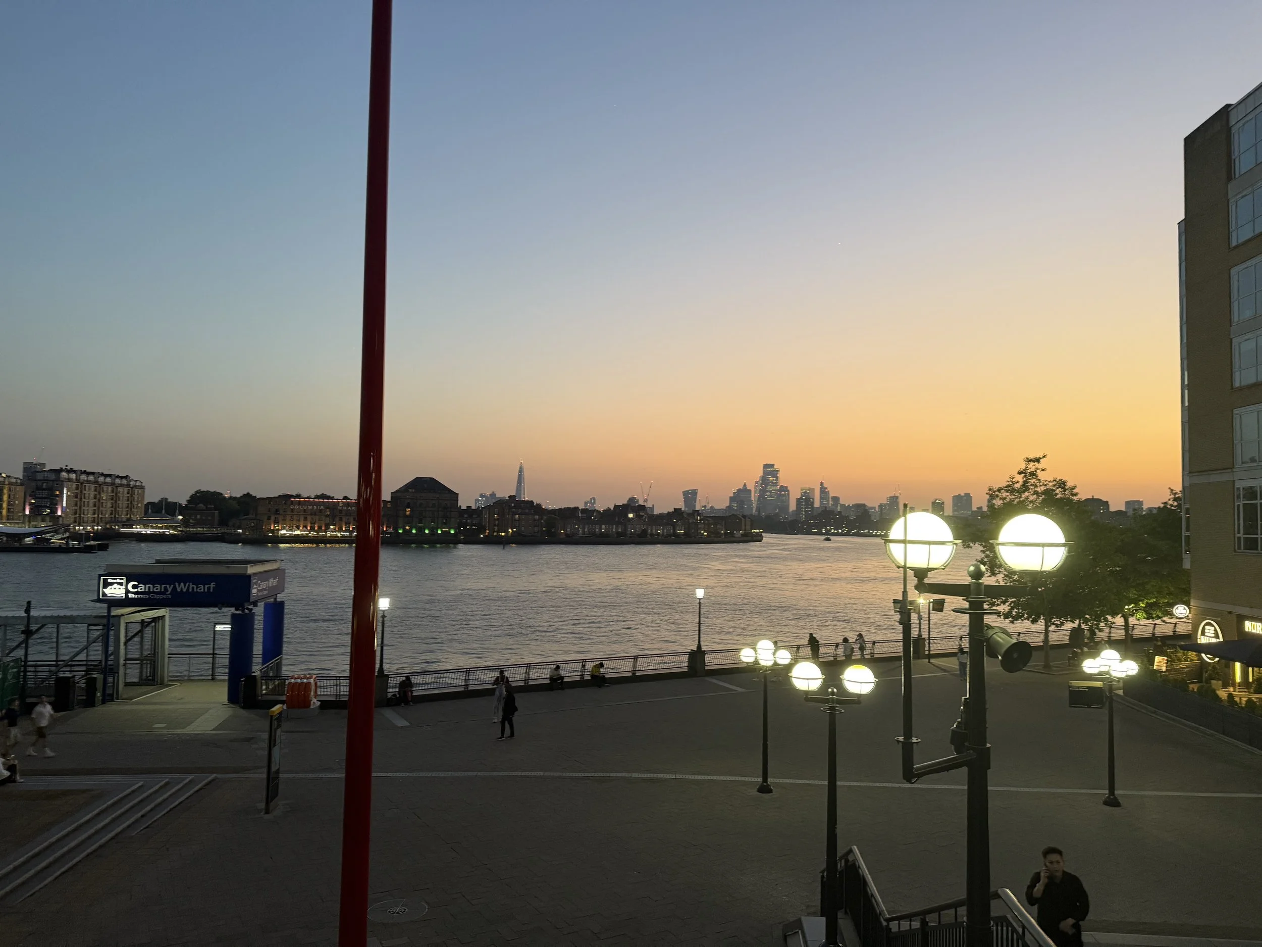 View of a city skyline at sunset, with buildings along the water, a prominent red flagpole, and a canal or river with a bridge in the foreground. City lights are beginning to turn on.