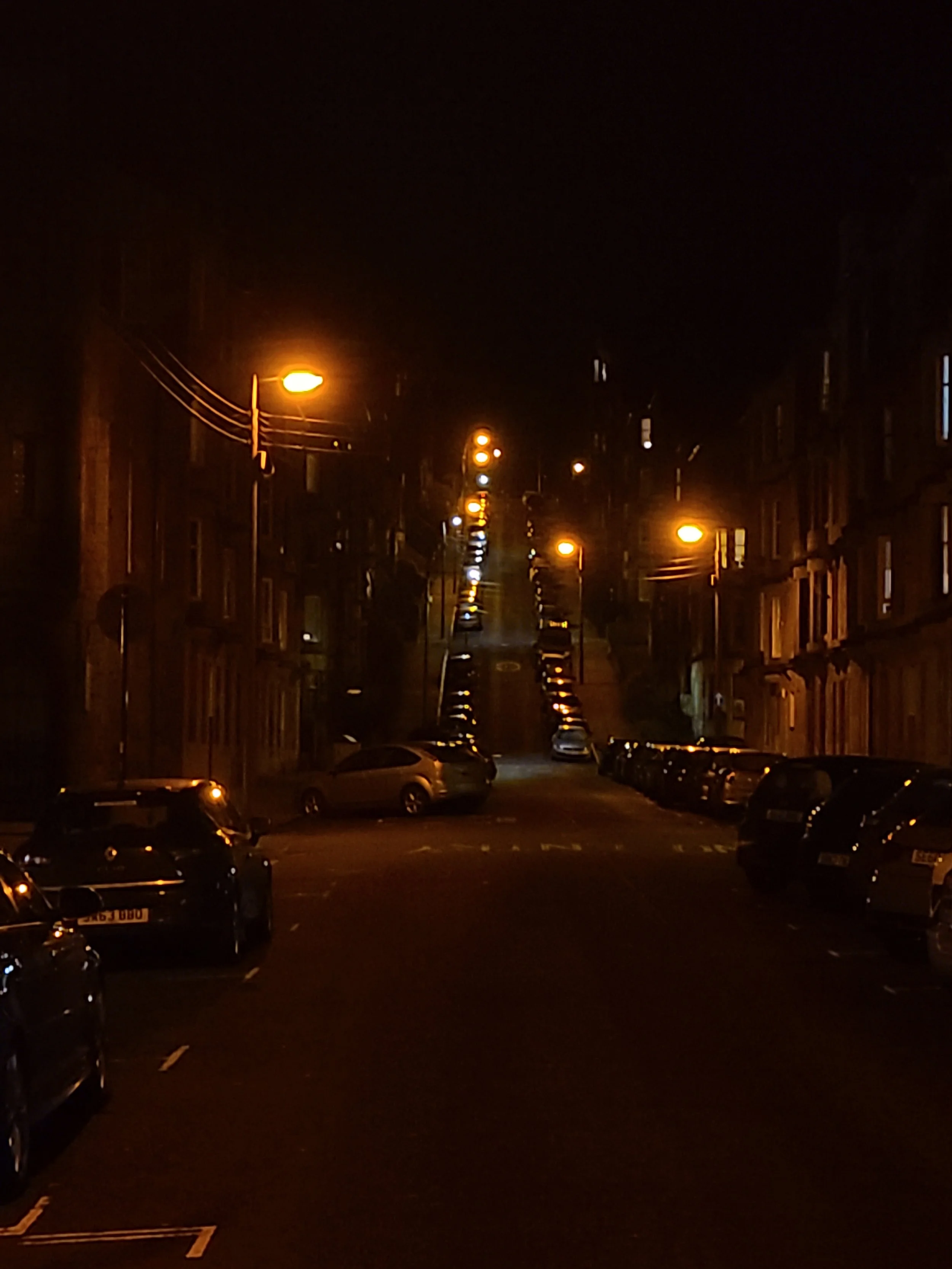 Nighttime city street scene with parked cars and streetlights illuminating the scene.