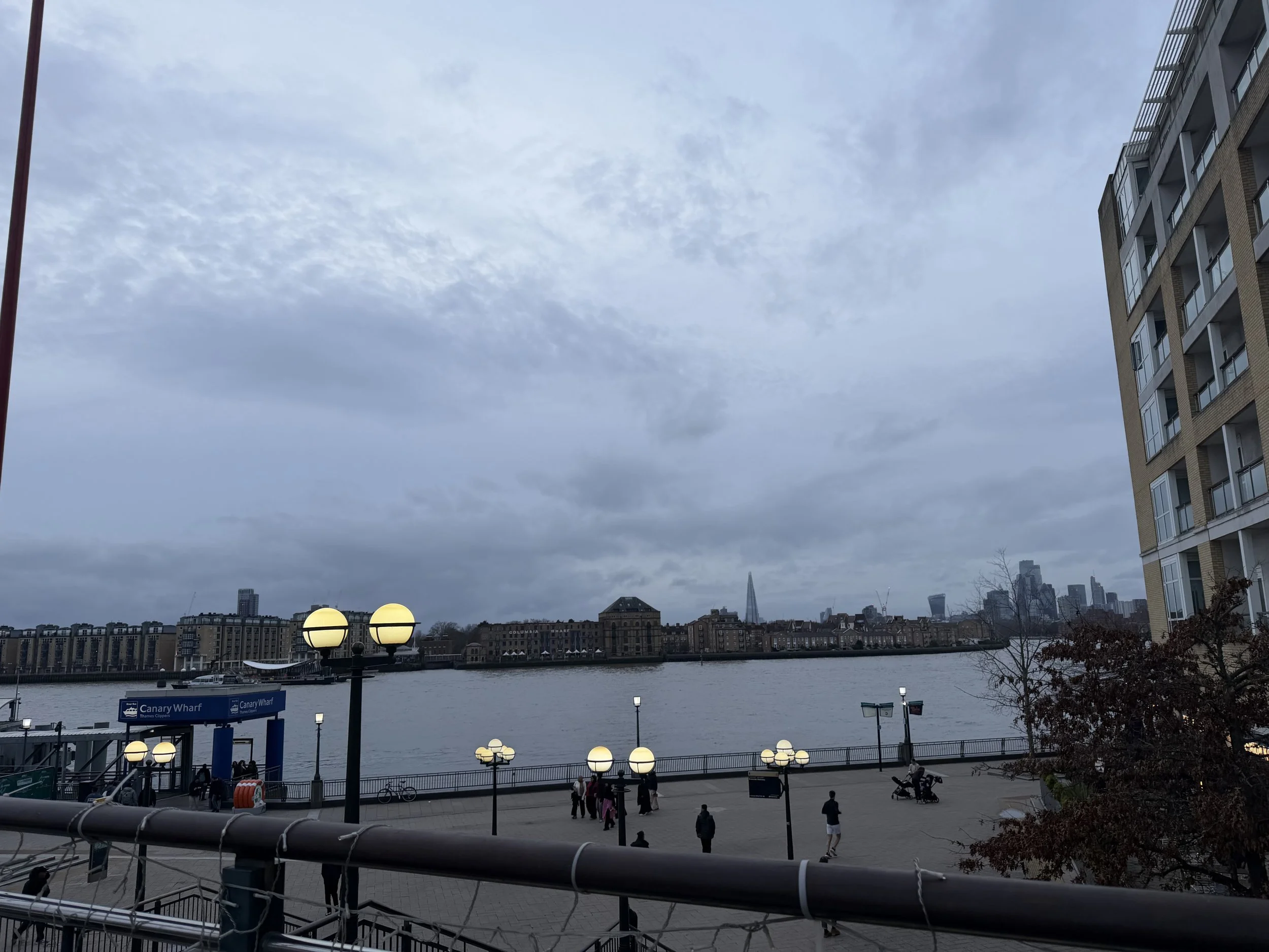 View of a city skyline across a river with modern buildings, a partly cloudy sky, and people walking near the waterfront.