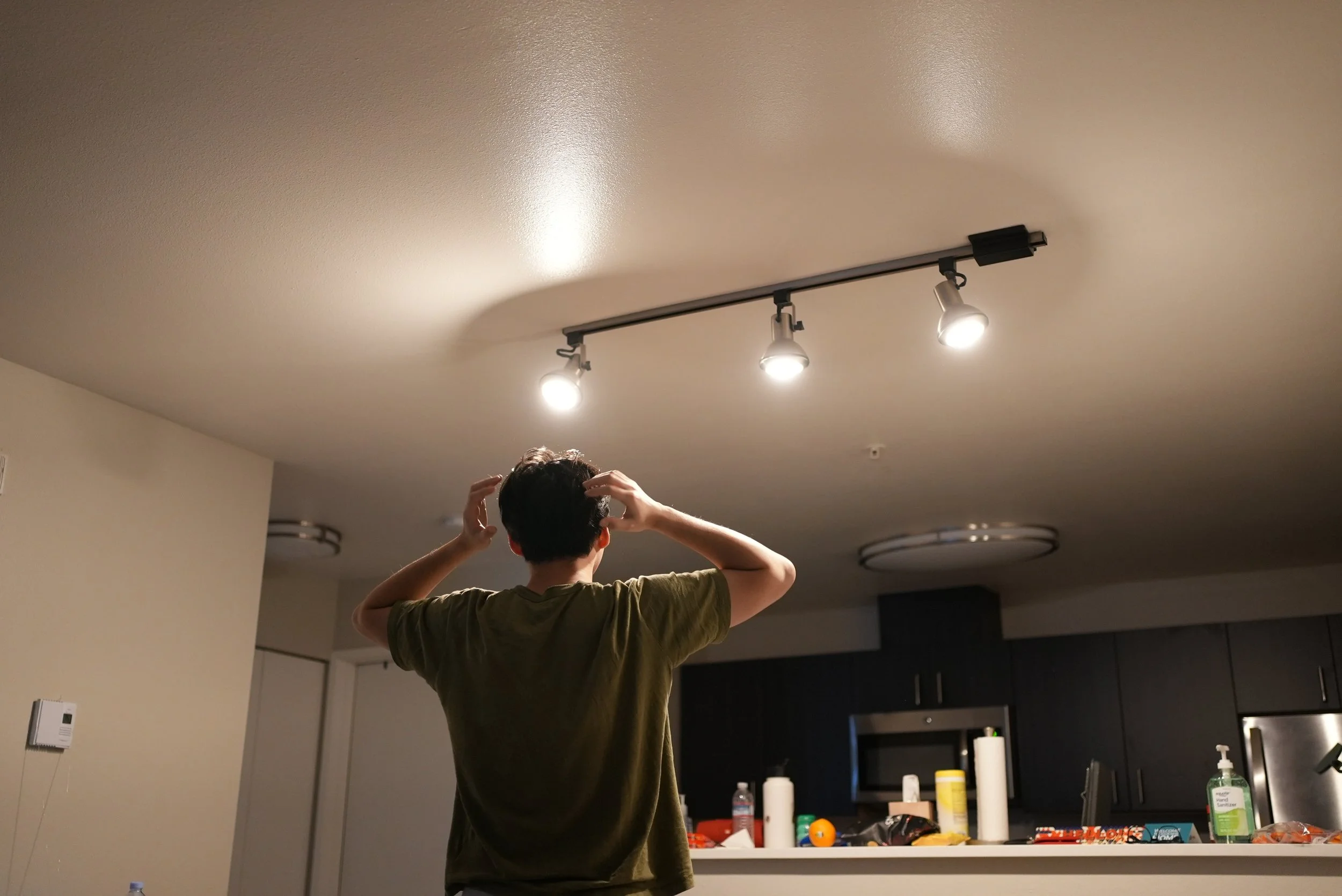 A person with dark hair wearing a green shirt, seen from behind, adjusting their hair in a room with a ceiling light, kitchen cabinets, and various objects on a counter.