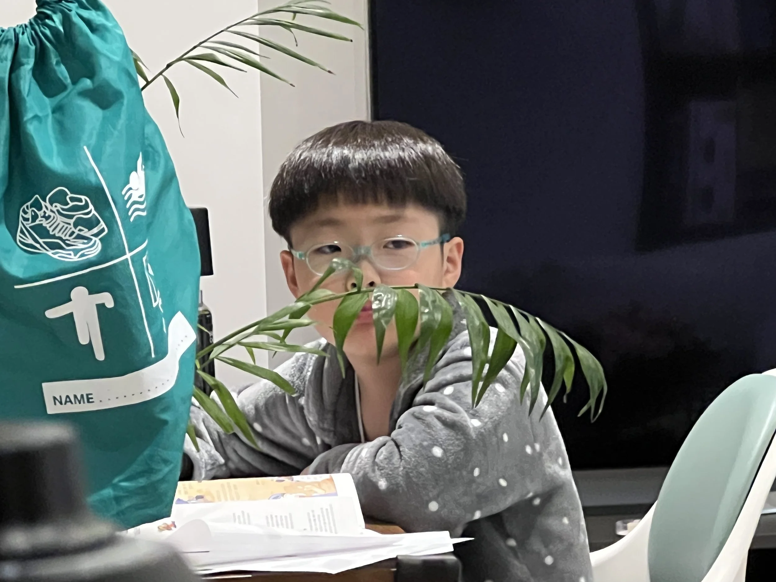 A young boy with glasses sitting at a table partially obscured by a green plant in the foreground. Behind him, part of a teal bag with a white graphic and a name label. Other background elements include a dark wall and a person in the background.