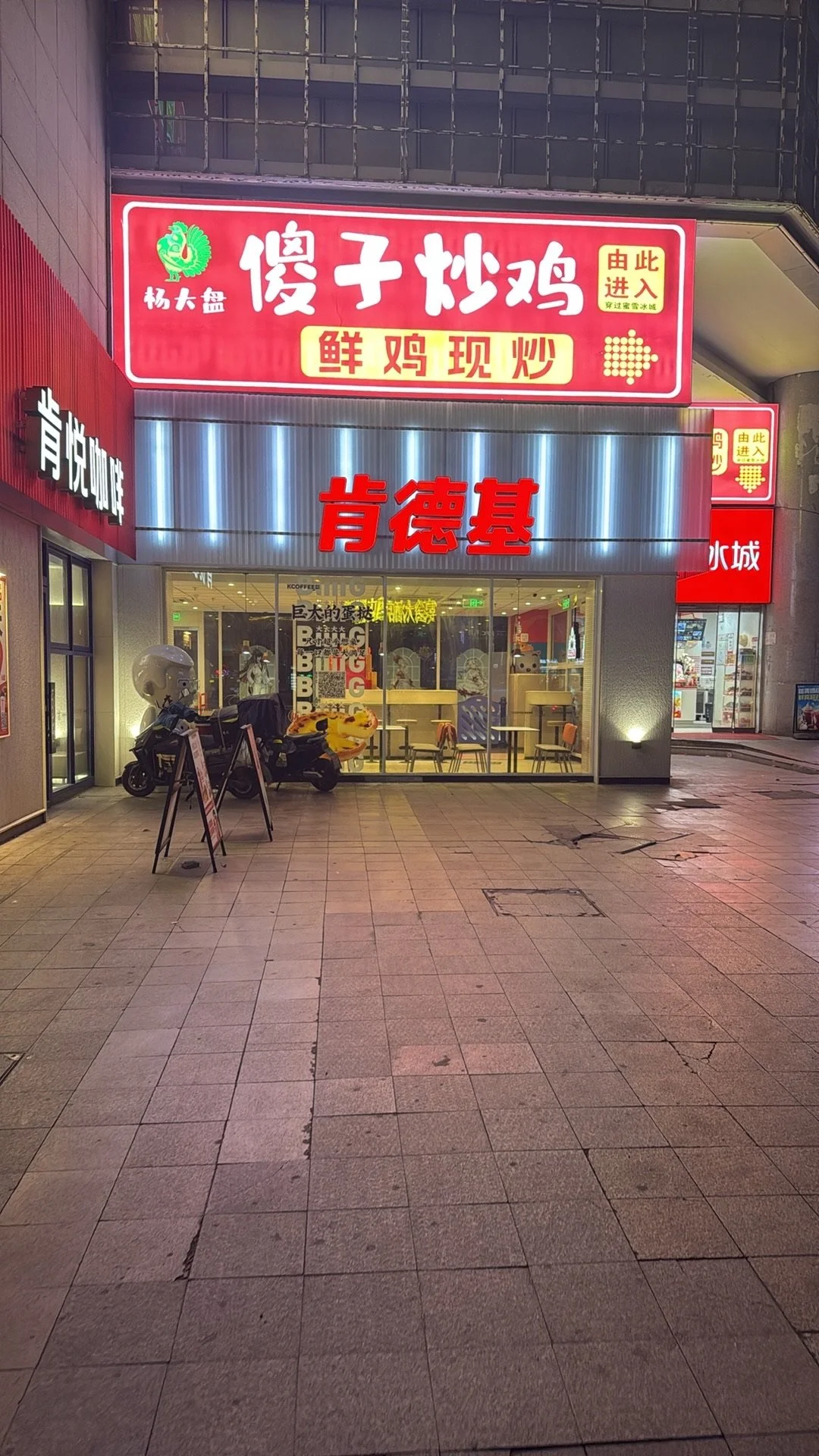 Night view of a restaurant with red illuminated signs in Chinese characters on the storefront. The inside of the restaurant is visible through large glass windows, showing tables and chairs. Outside, there is a scooter parked near the entrance and a 