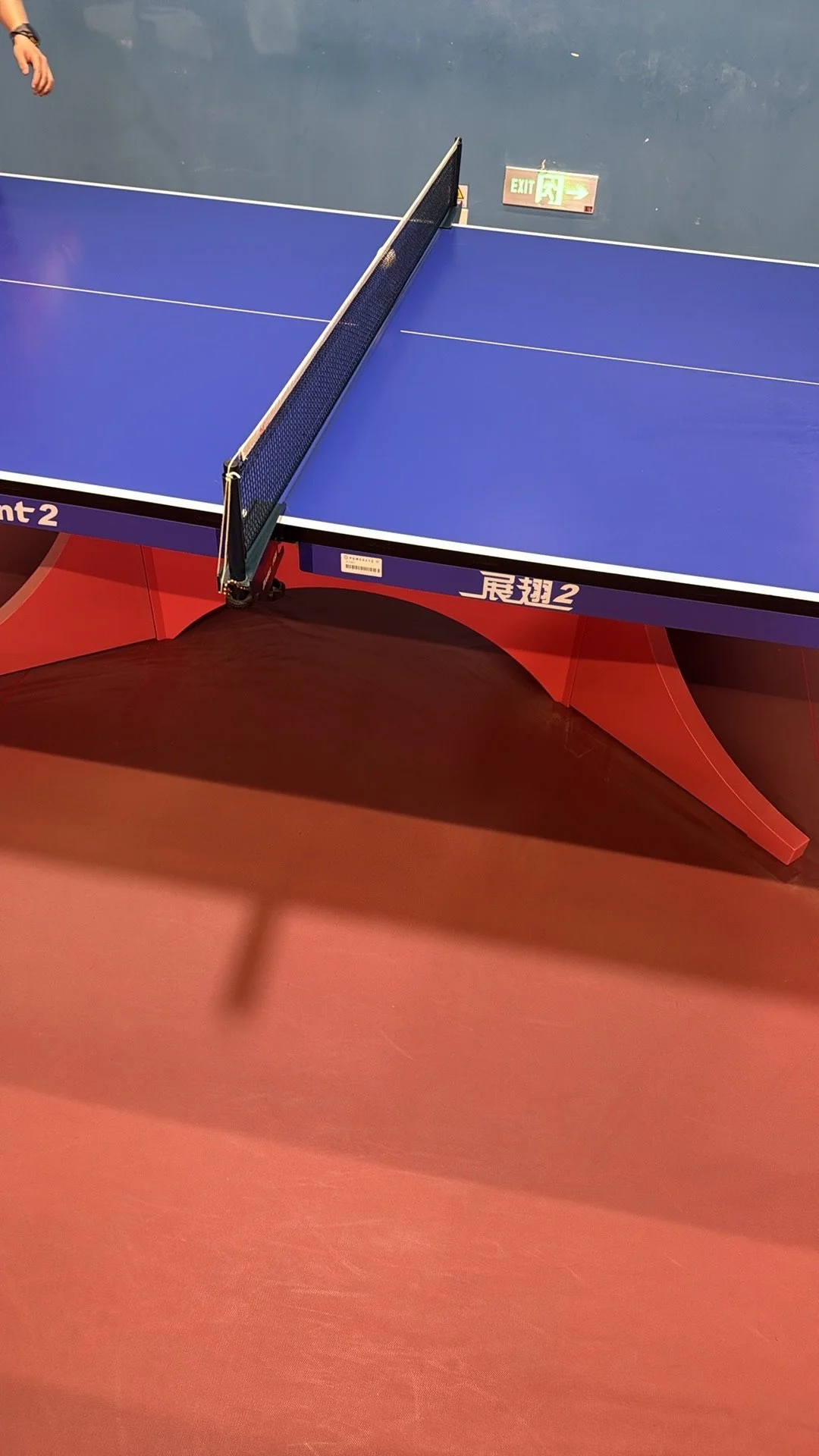 An indoor blue ping pong table with orange legs and a black net, on a red floor, with an exit sign on the wall in the background.