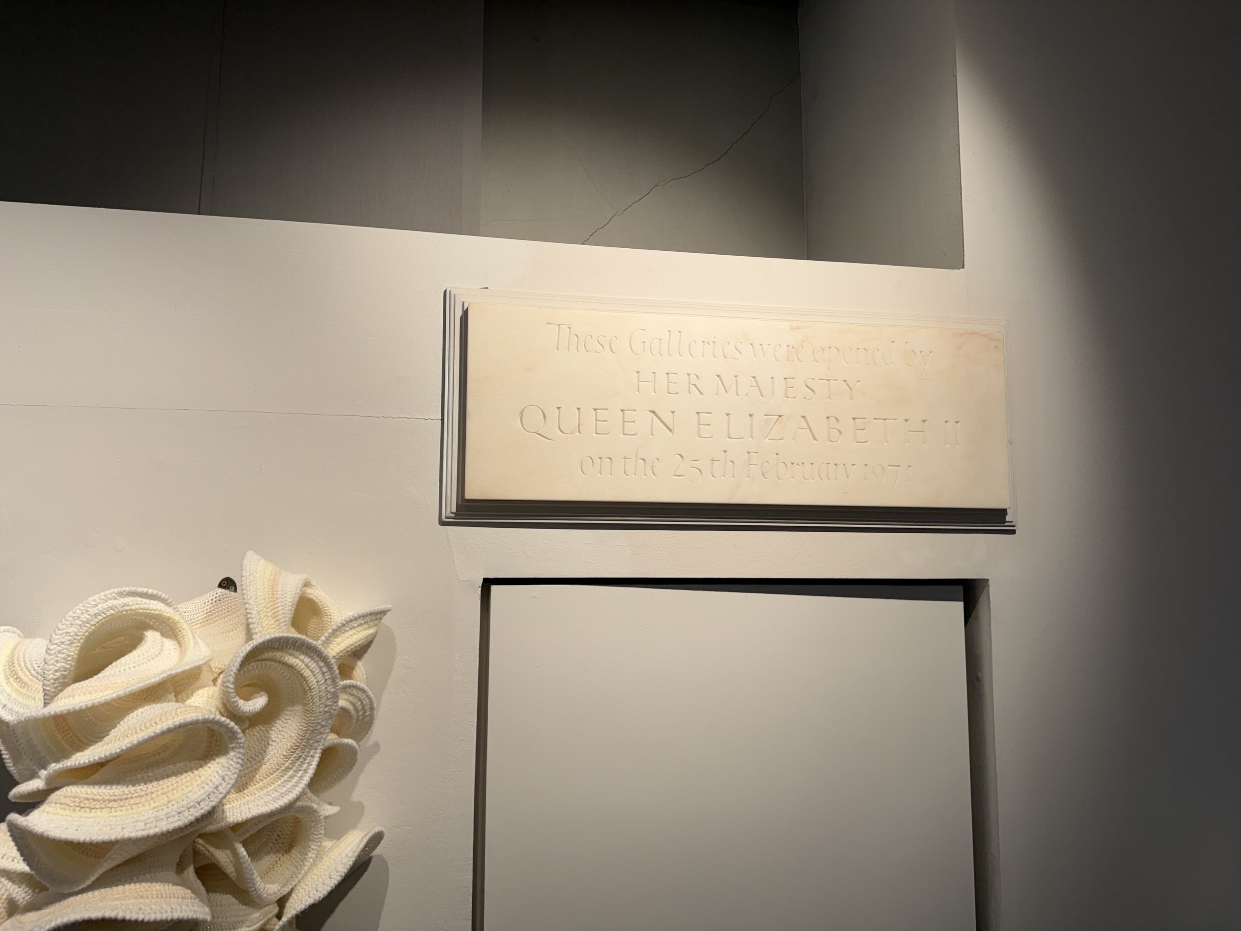A beige floral sculpture with intricate, layered petals on the left side and a marble plaque with text on the right side, mounted on a white wall.