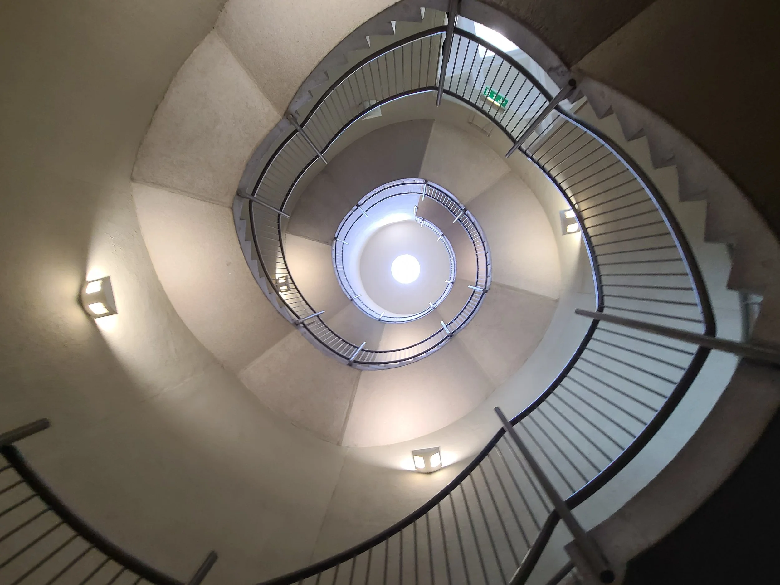 Looking down a spiral staircase with metal railings and stairs, illuminated by wall-mounted lights along the beige walls, with a bright circular light source at the bottom.