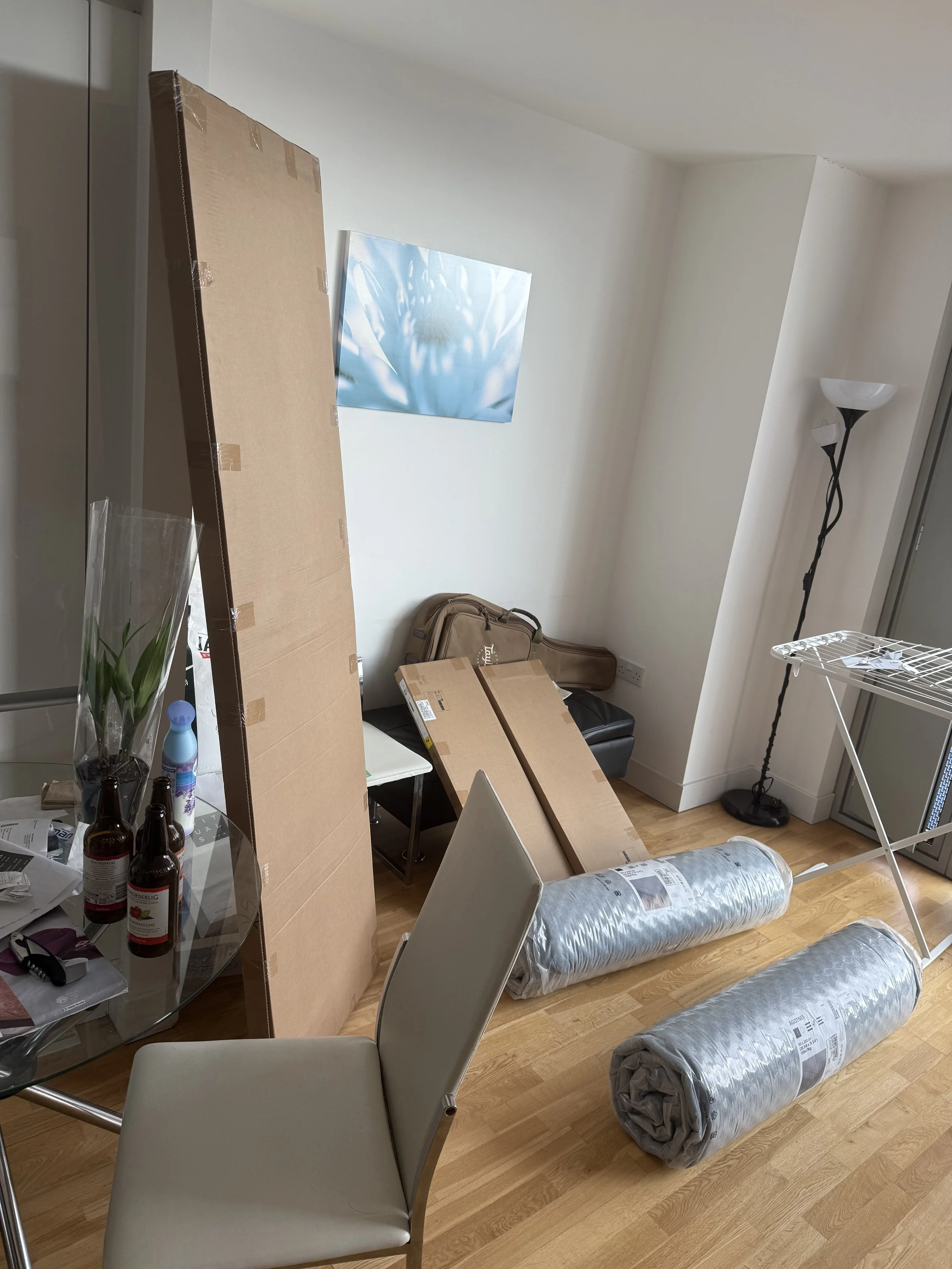 Living room with cardboard boxes, rolled-up mattresses, a table, chairs, a floor lamp, and a colorful artwork on the wall.