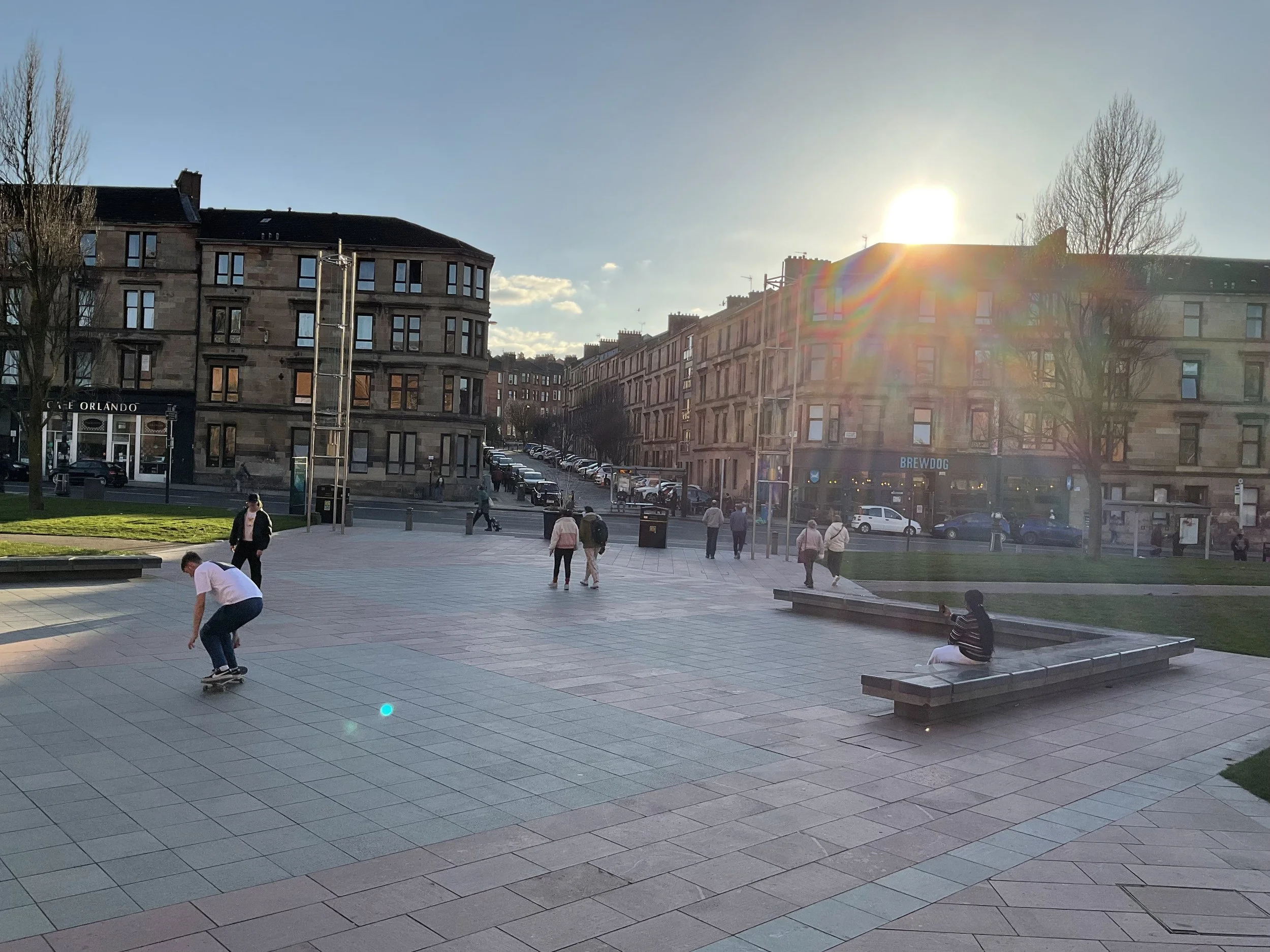 A city street scene in the late afternoon or early evening with a blue sky and sunlight creating a rainbow lens flare. People are walking and skateboarding on a paved public square, with parked cars along the street and multi-story buildings in the b
