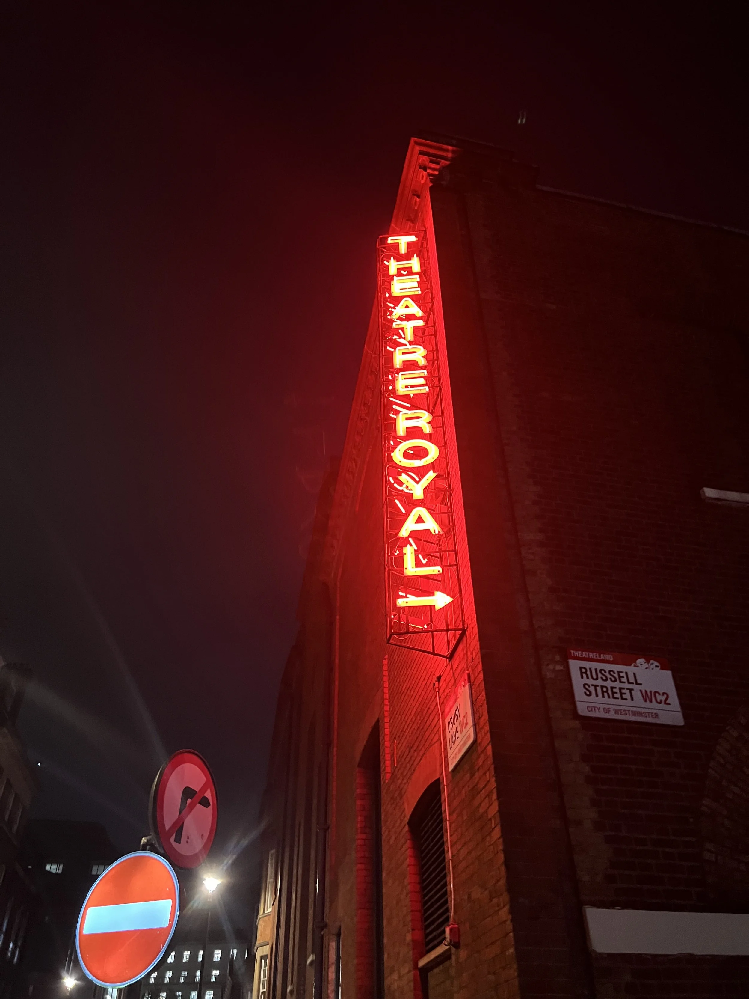 Nighttime photo of a tall red neon sign that reads "VAULTS & PARTY" attached to a brick building. There are street signs including a no entry sign and a no parking sign, and a street name sign that reads "RUSSELL STREET WC2" in Westminster.