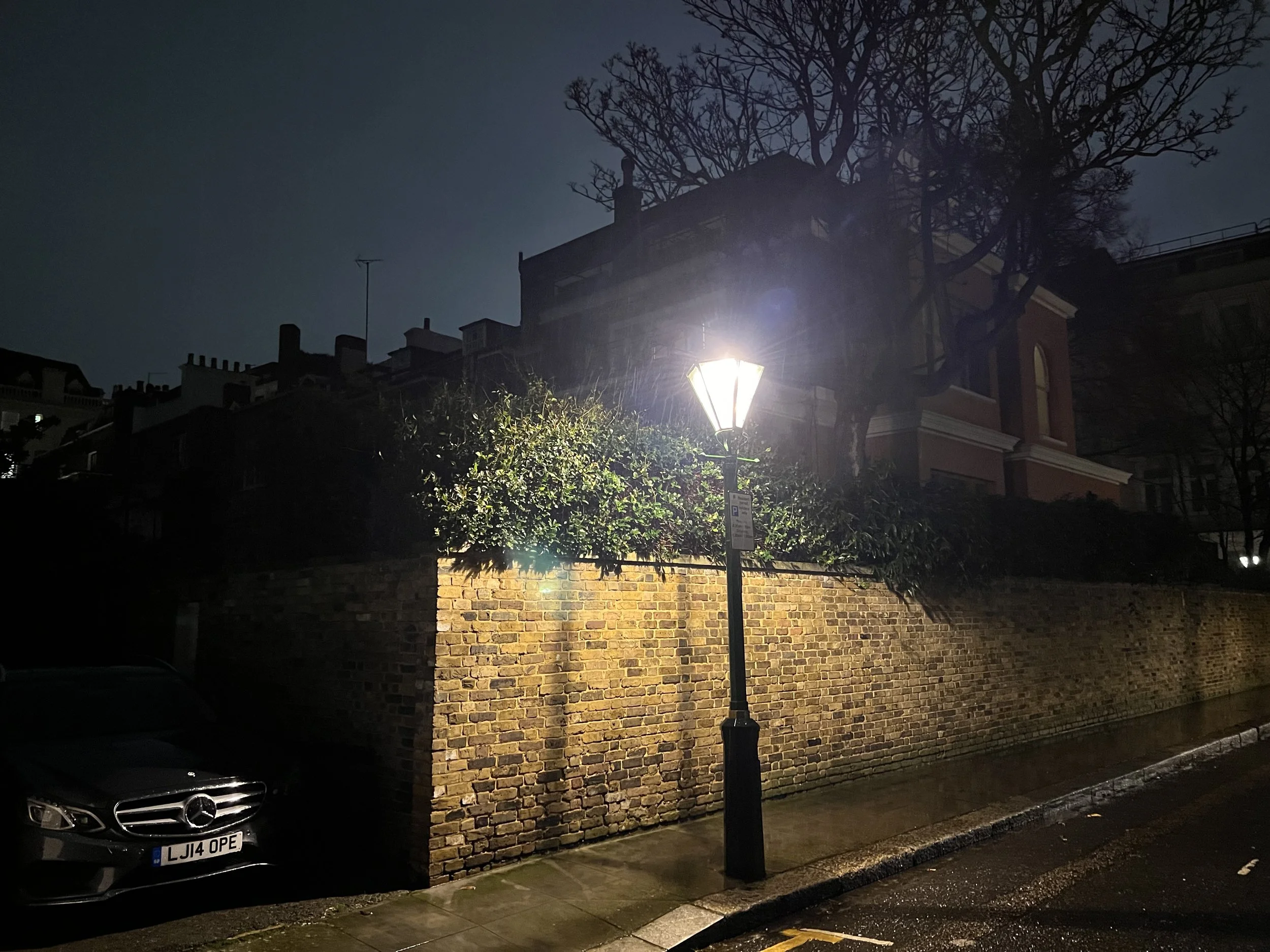 Nighttime street scene with a brick wall, a street lamp, bare tree, and a parked black Mercedes-Benz car.