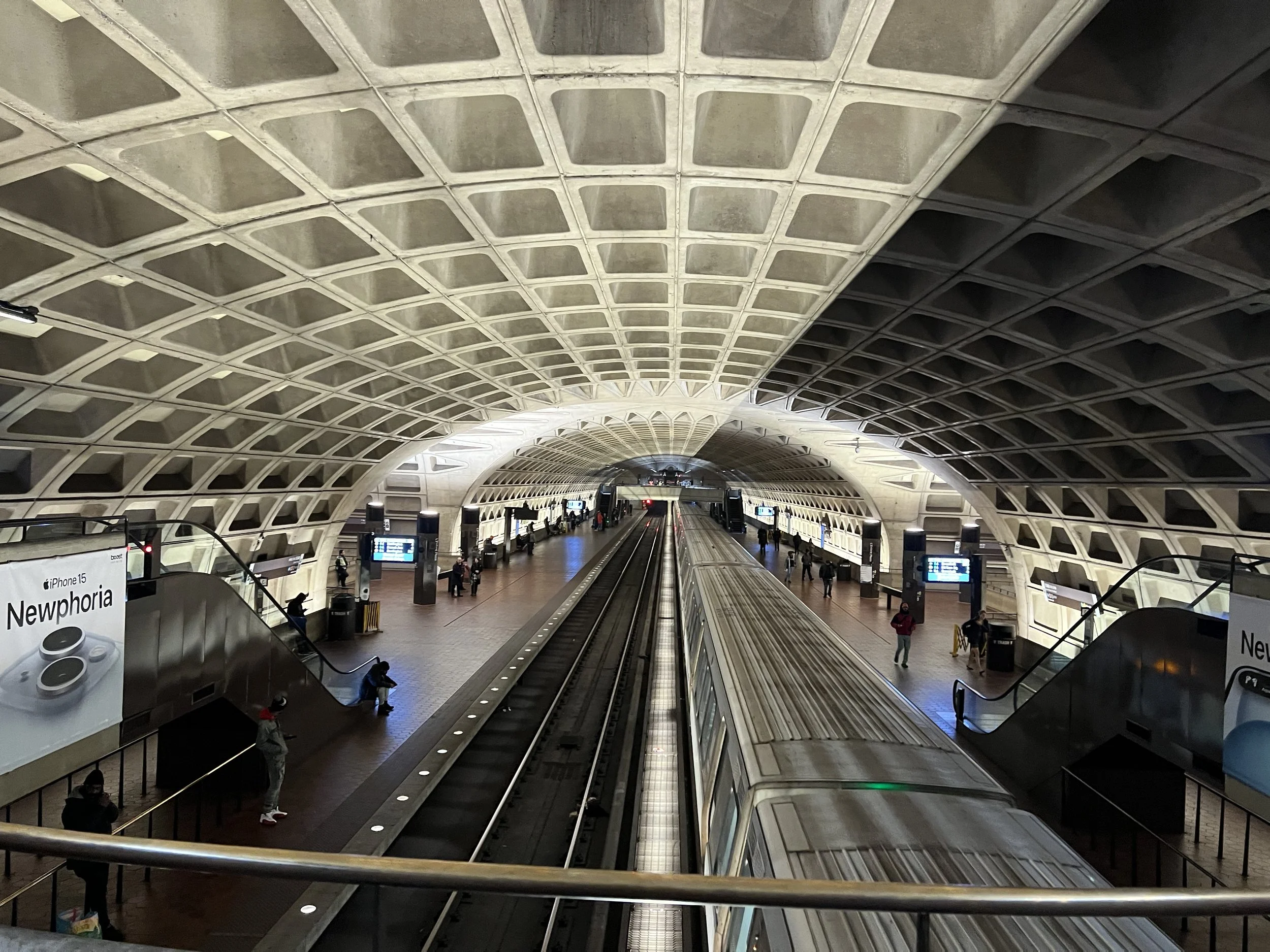 Interior view of a modern subway station with an arched ceiling featuring a grid design. There are escalators on both sides and a moving walkway in the center, with several people walking or sitting around. Digital screens and advertisements are visi