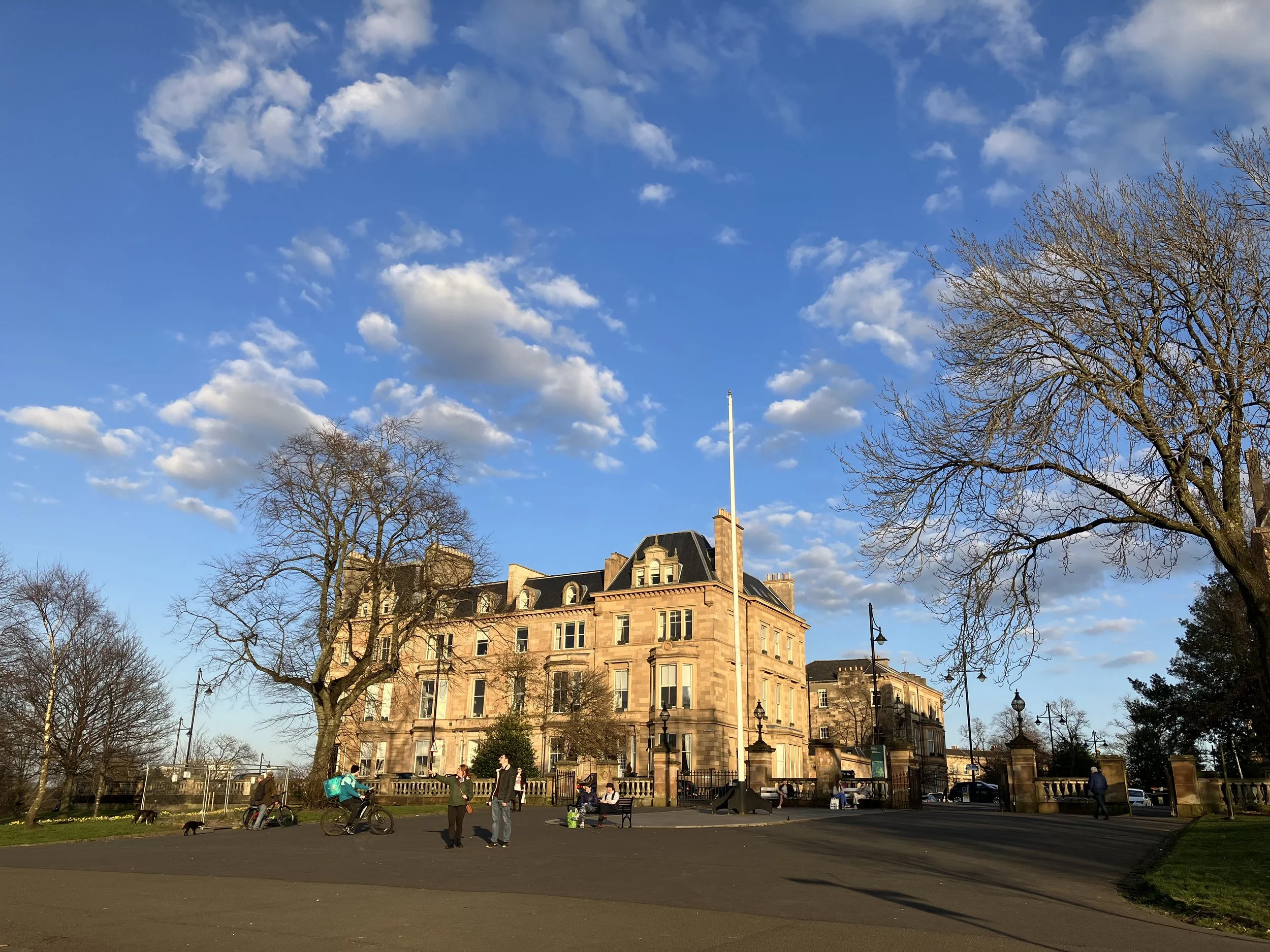 A storefront building with multiple windows and ornate architectural details, surrounded by leafless trees, with a park and people in the foreground under a blue sky with scattered clouds.