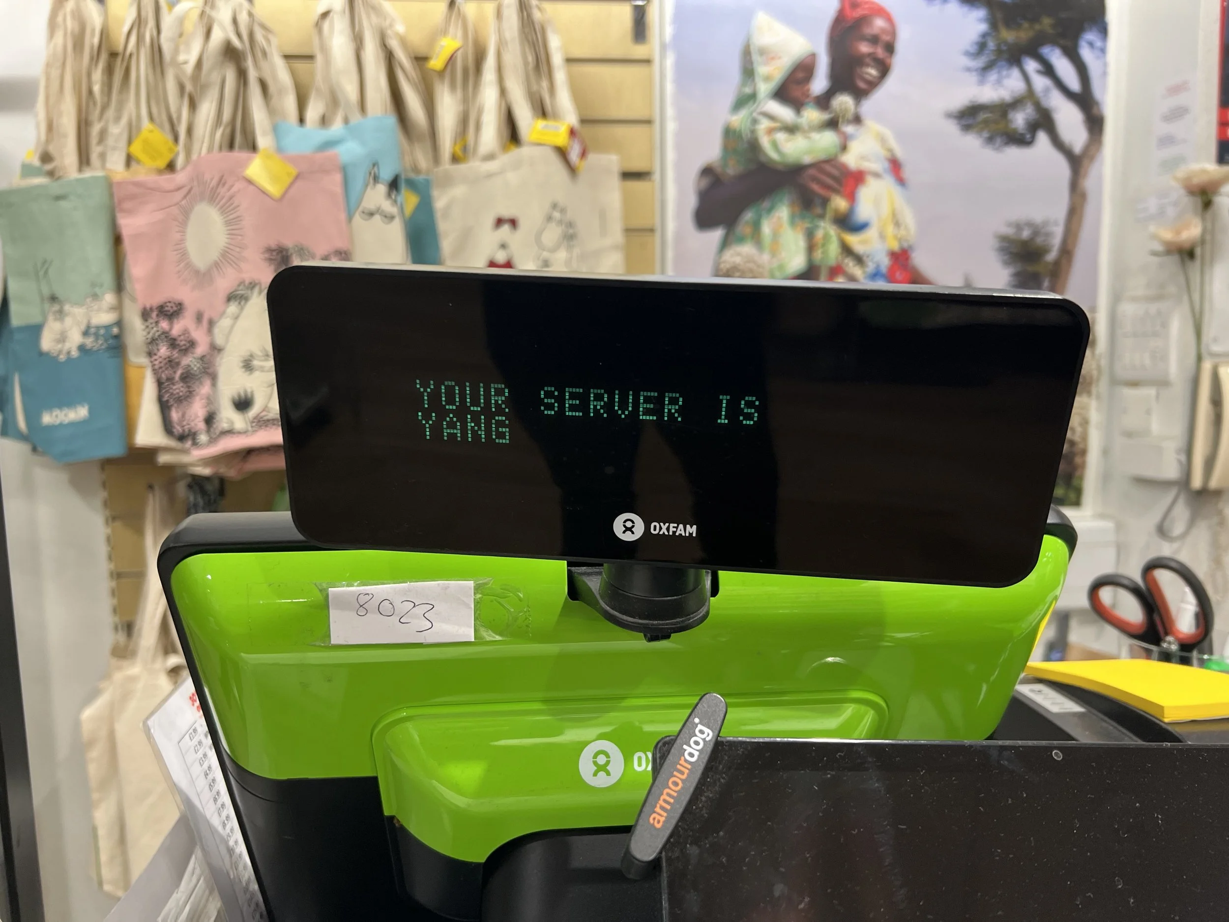 A checkout counter at a store with a digital display that reads 'YOUR SERVER IS YANG'. In the background, there are shopping bags with various designs hanging on a wall.