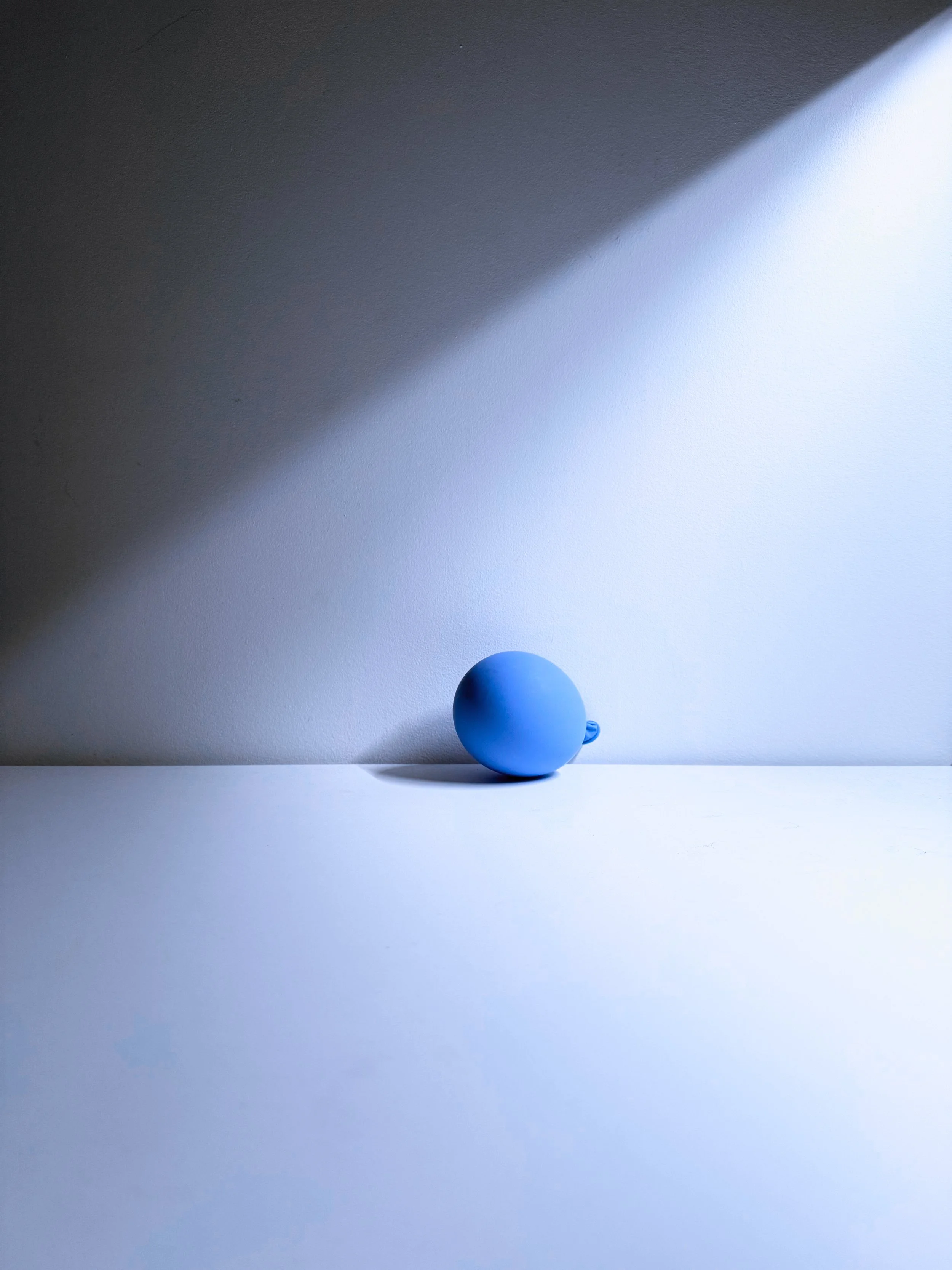 A small blue balloon resting on a white surface. Shadows and light create a gradient on the background wall.