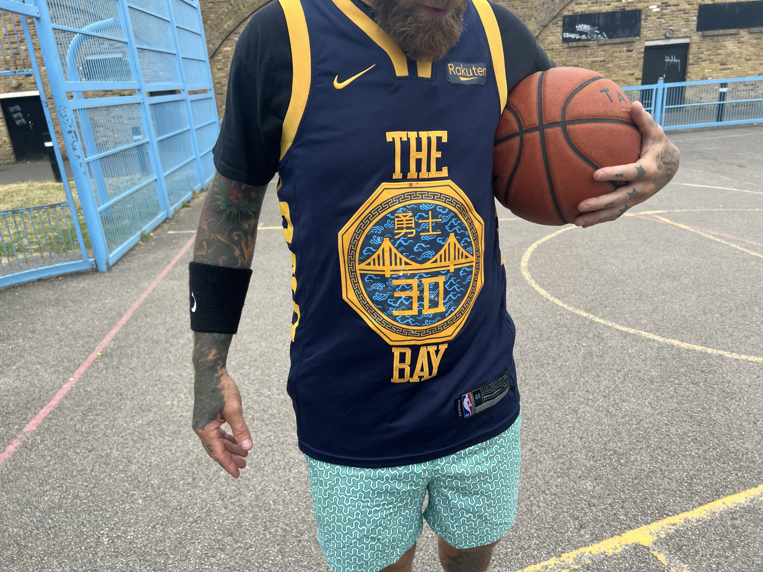 Person holding a basketball on an outdoor basketball court, wearing a blue and yellow basketball jersey with the words "THE BAY" and a large logo, and patterned shorts.