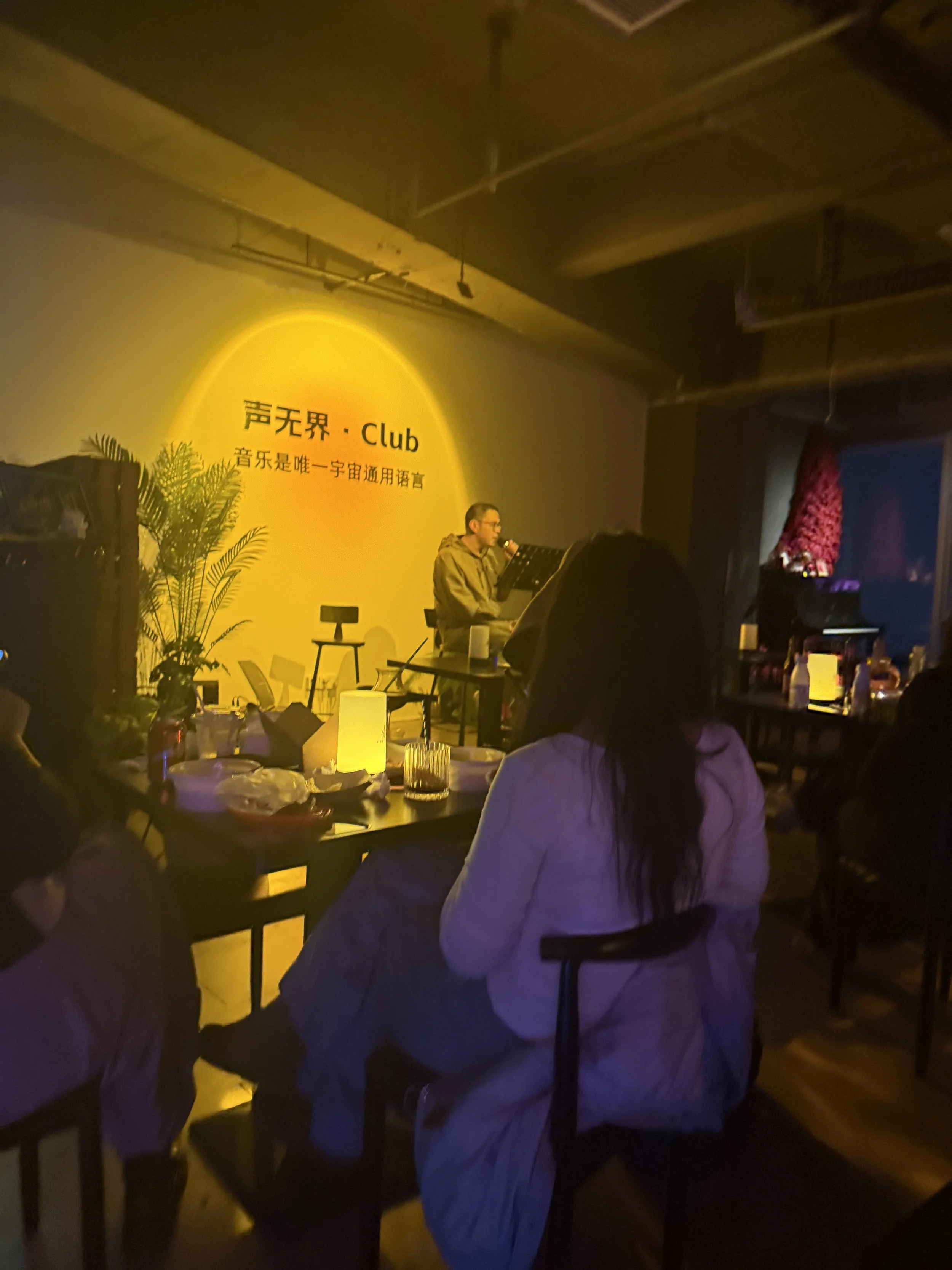 A man singing into a microphone on stage at a club with a yellow wall that has Chinese characters and the words 'Club', surrounded by people sitting at tables with drinks.